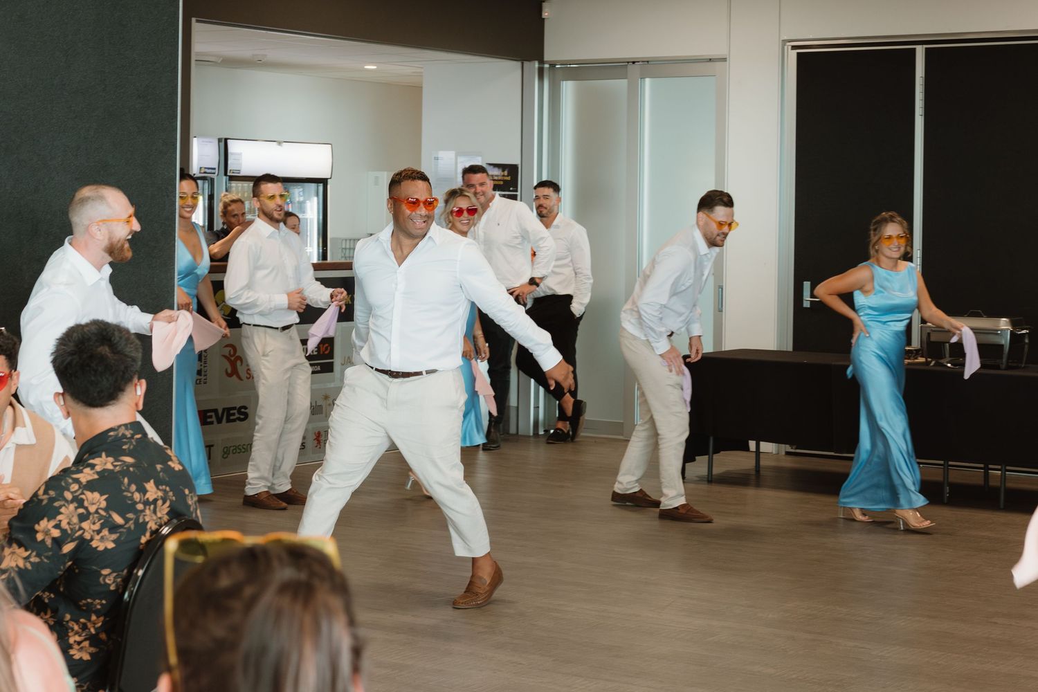 Group of people dancing at a wedding reception in a modern venue, men in white shirts and woman in blue gown.