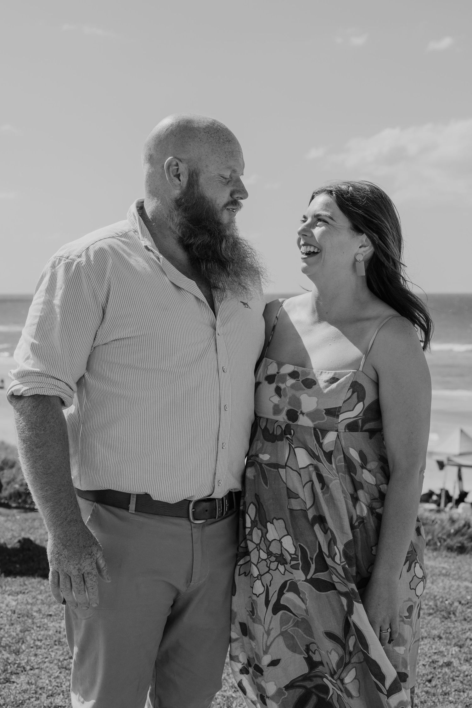 Black and white photo of a bearded man and laughing woman sharing a joyful moment on a beach.