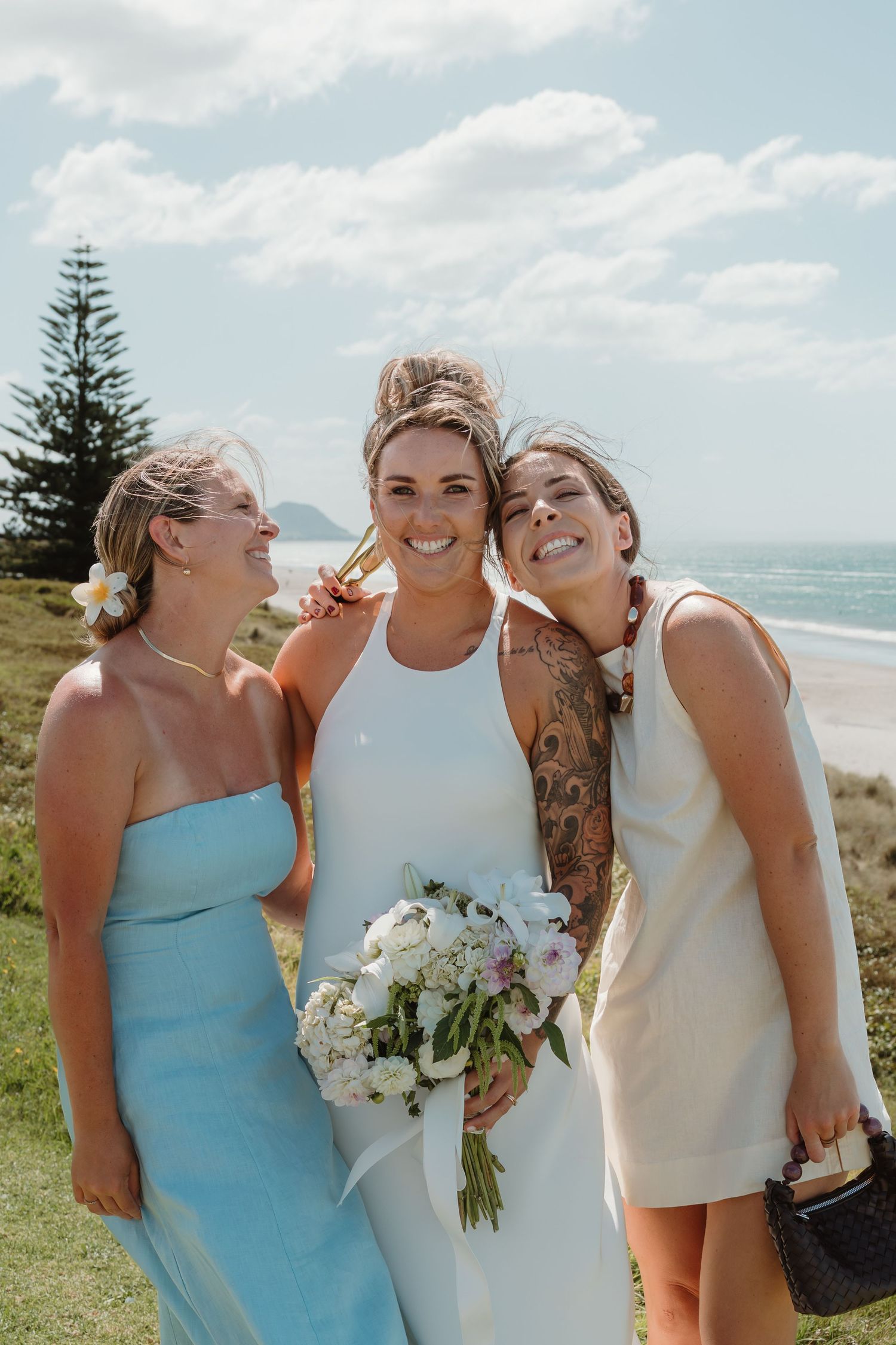Bride in white halter dress holding bouquet poses with two bridesmaids at a scenic beach wedding in New Zealand.