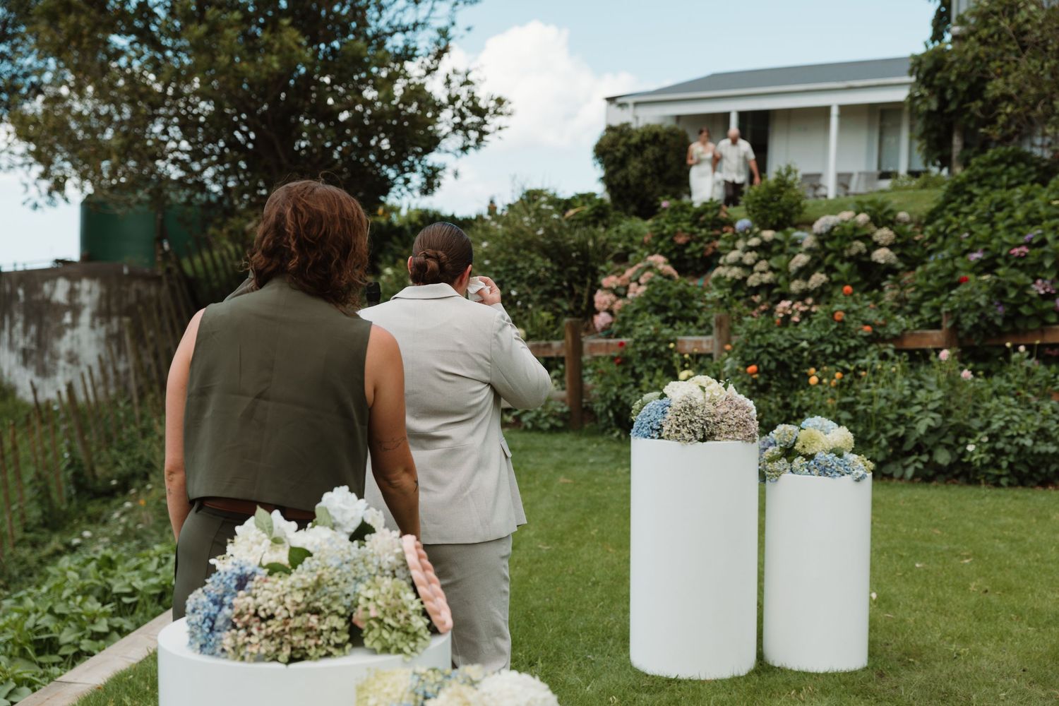 Two guests watch an outdoor garden wedding ceremony with white pedestals adorned with blue and white hydrangea floral arrangements.
