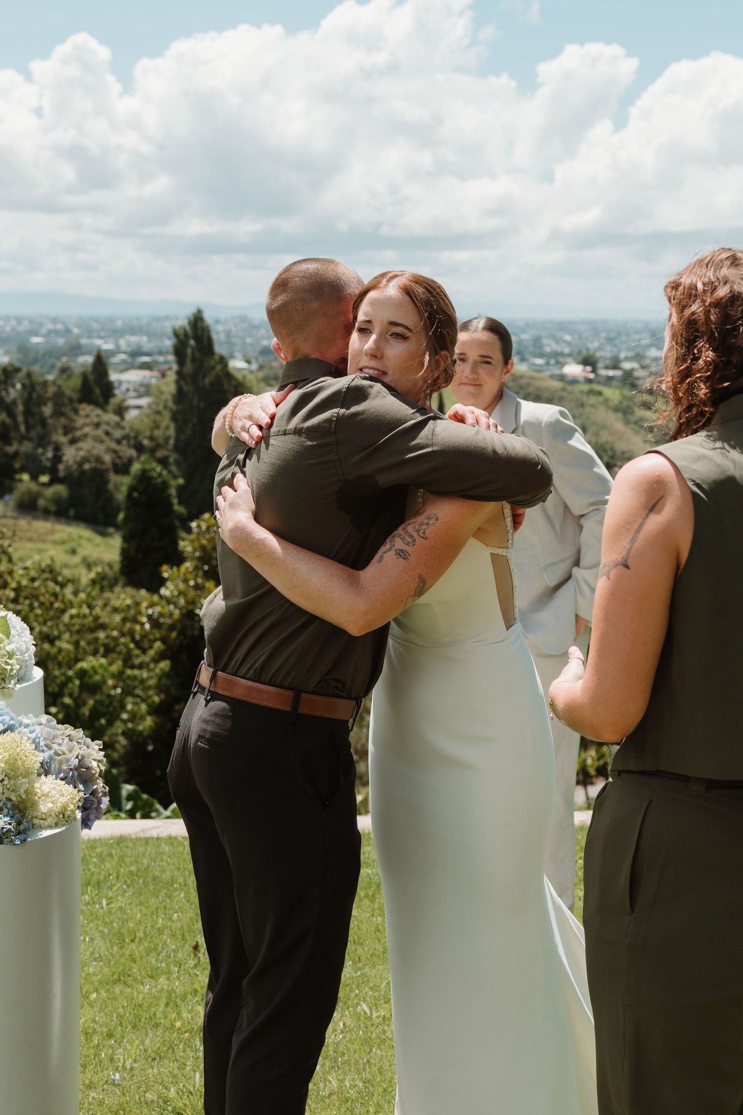 Bride in white dress hugging guest at outdoor wedding ceremony with scenic landscape backdrop.