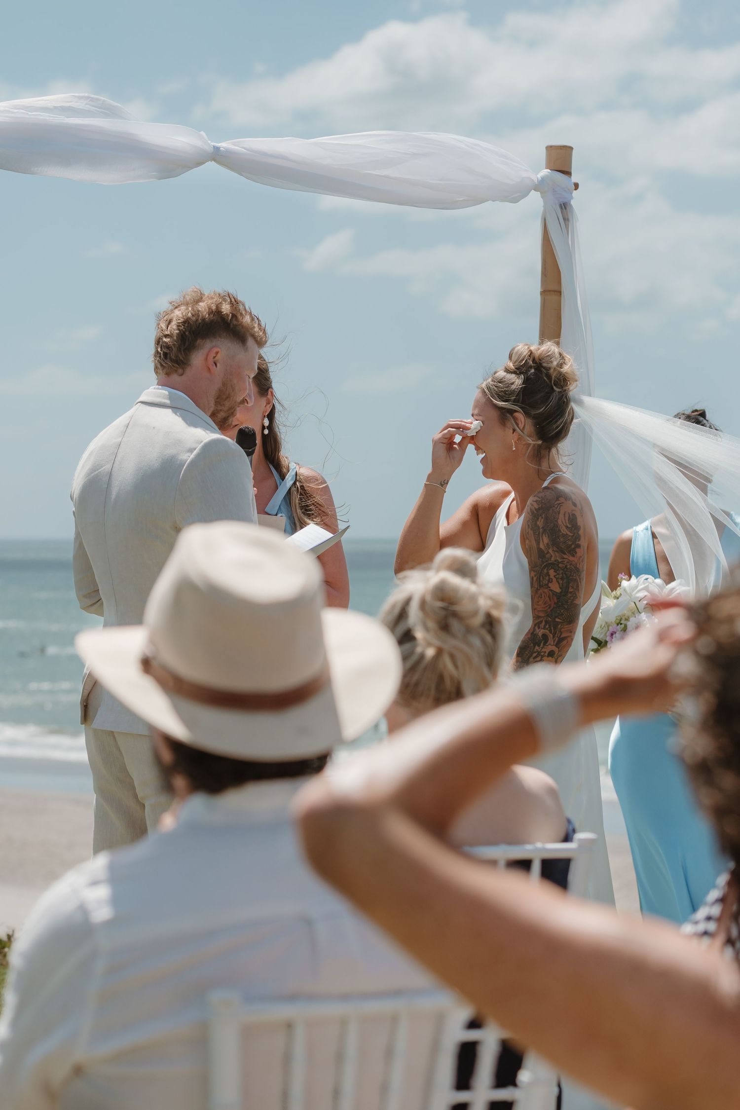 Couple exchanging vows at a romantic beach wedding ceremony under a white draped bamboo arch.