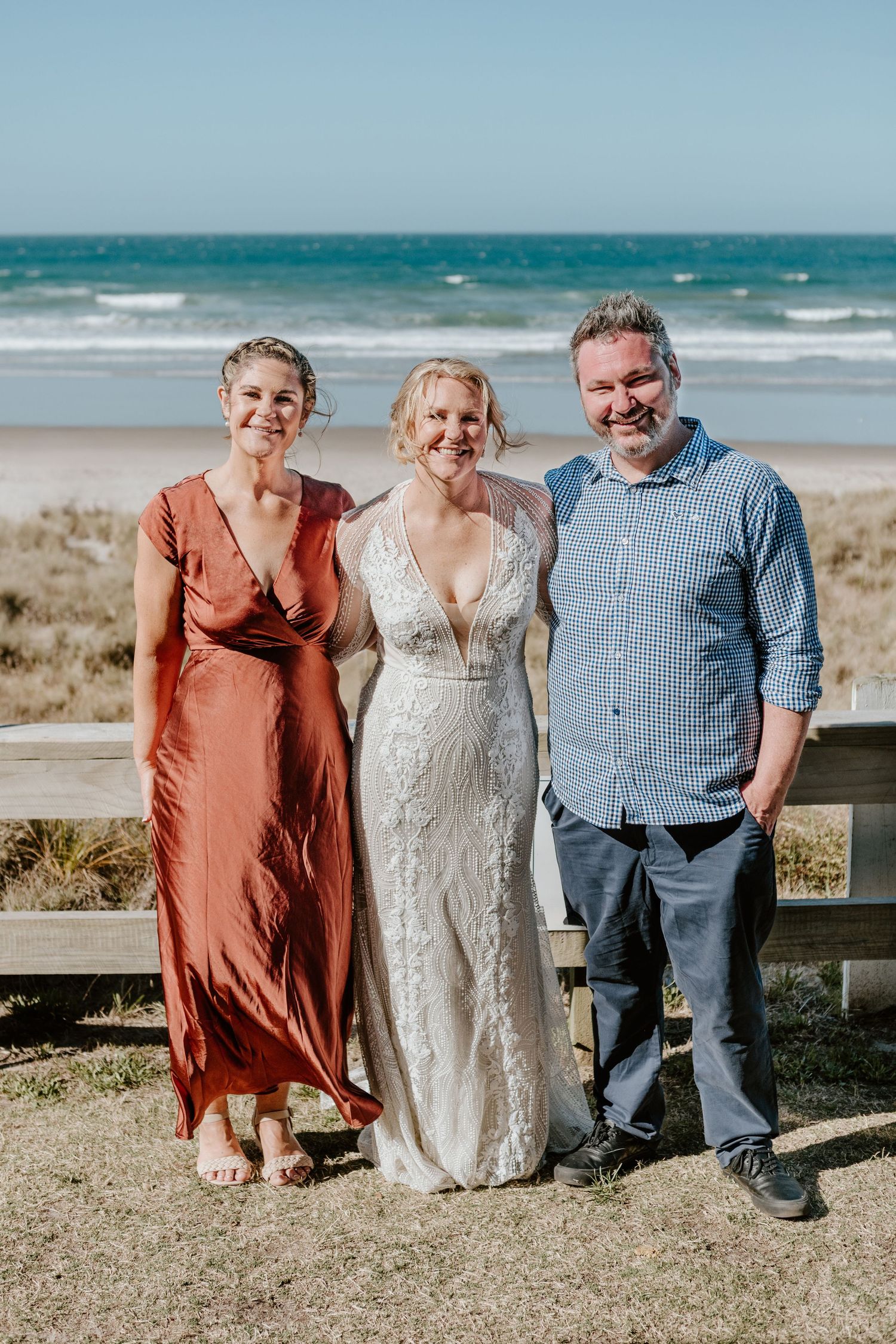 Small wedding group stands together on beach boardwalk with ocean waves in background. Papamoa Surf Lifesaving Club Wedding