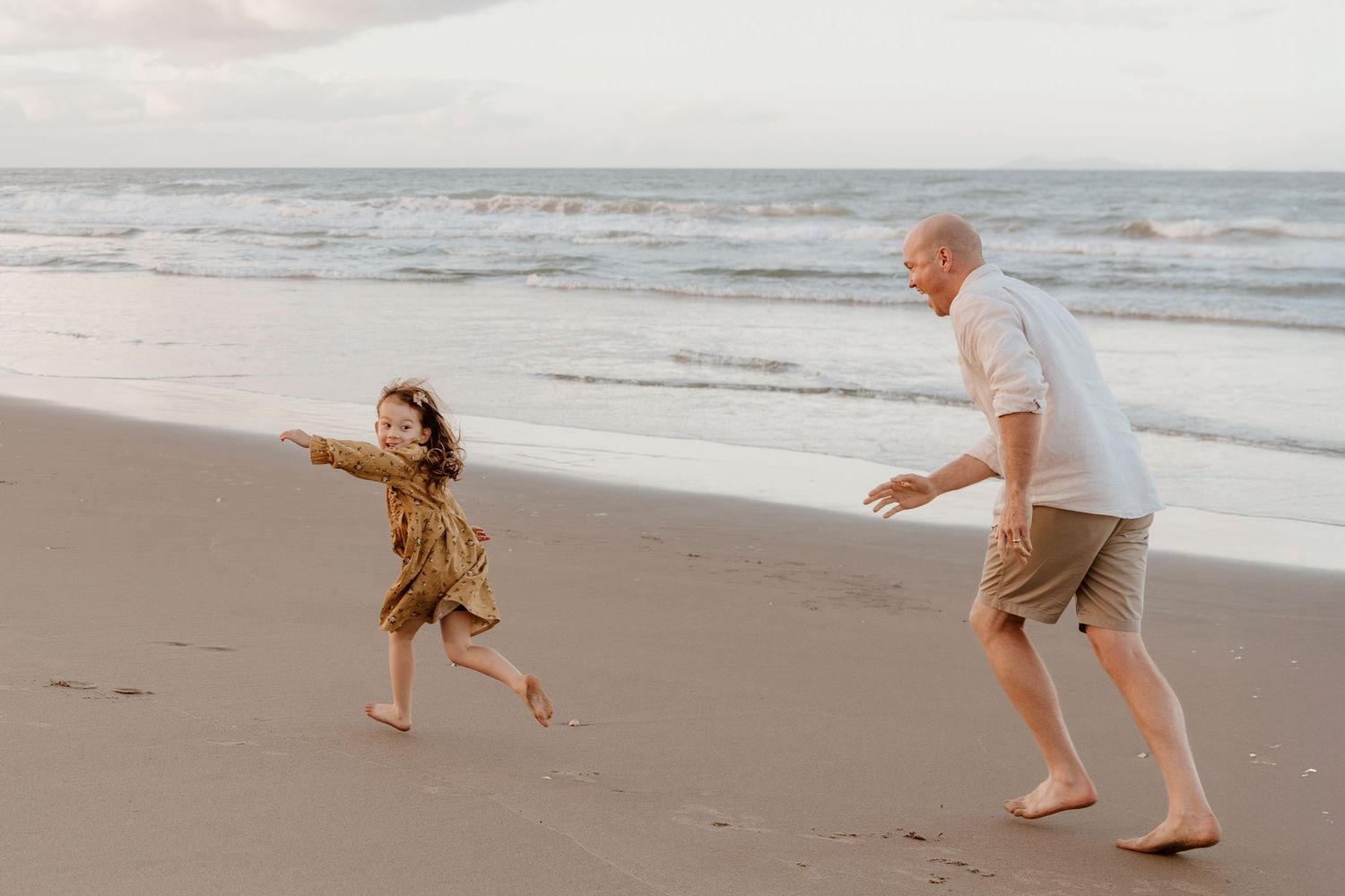 A playful chase scene between dad and daughter running barefoot along a serene beach at sunset. Papamoa, Tauranga family photographer, fun