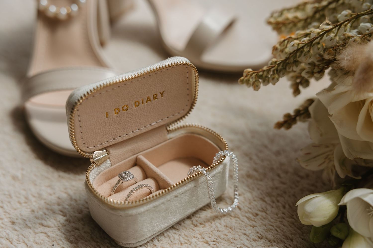 Elegant wedding accessories including pearl necklace, nude heels, and delicate floral details arranged on cream fabric.