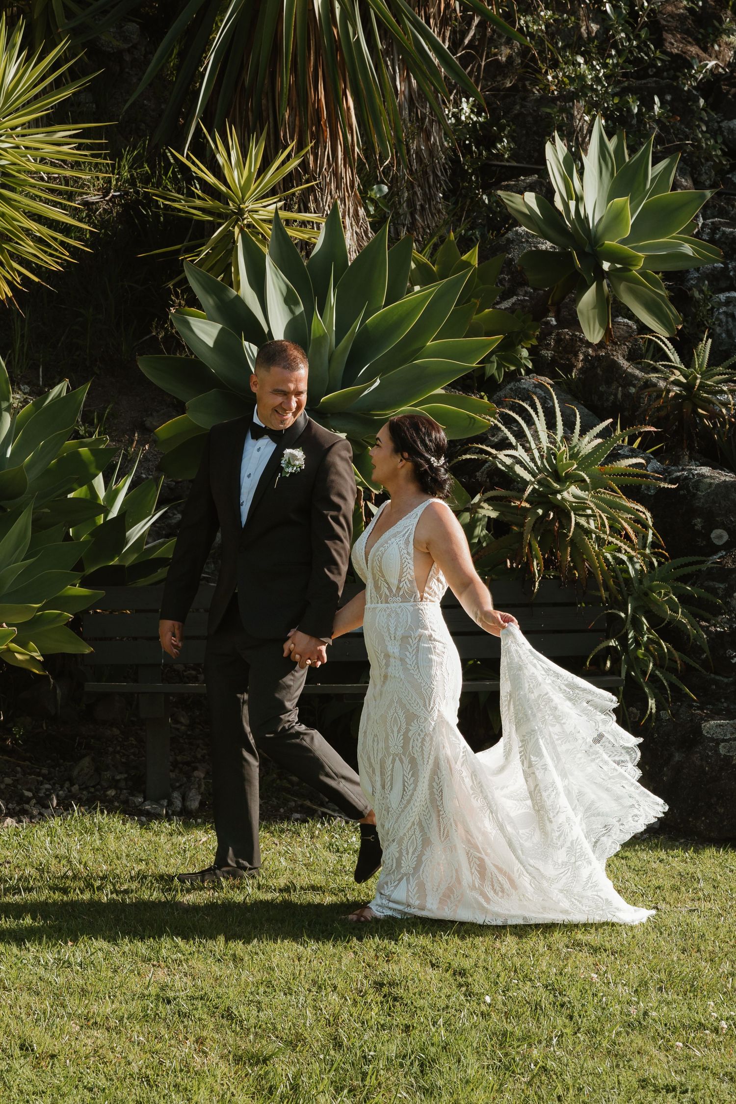 A couple in wedding attire dances together on a lush green lawn surrounded by tropical plants and palm trees.