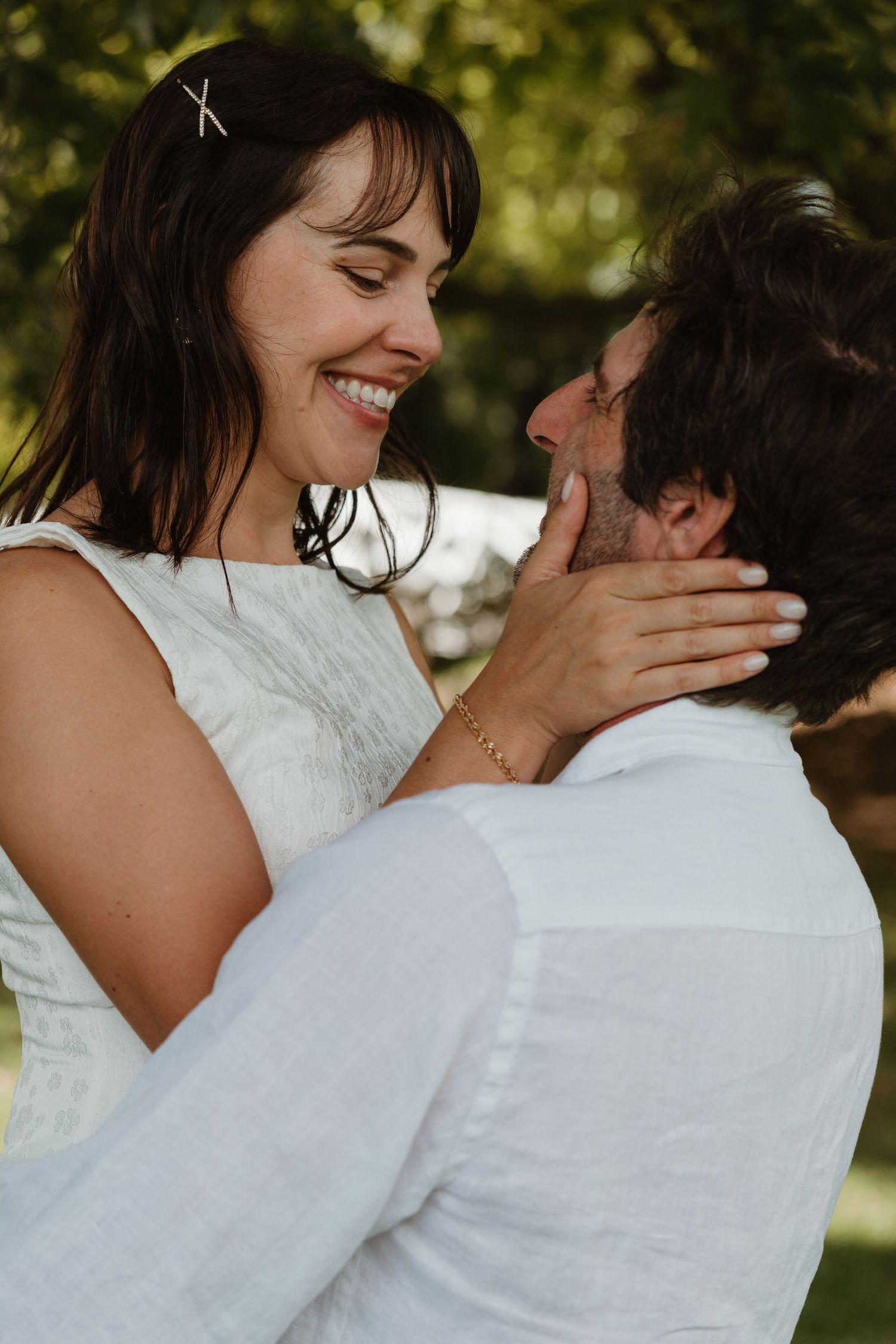 A smiling bride in white holds her groom's face tenderly outdoors, sharing a romantic moment surrounded by greenery.