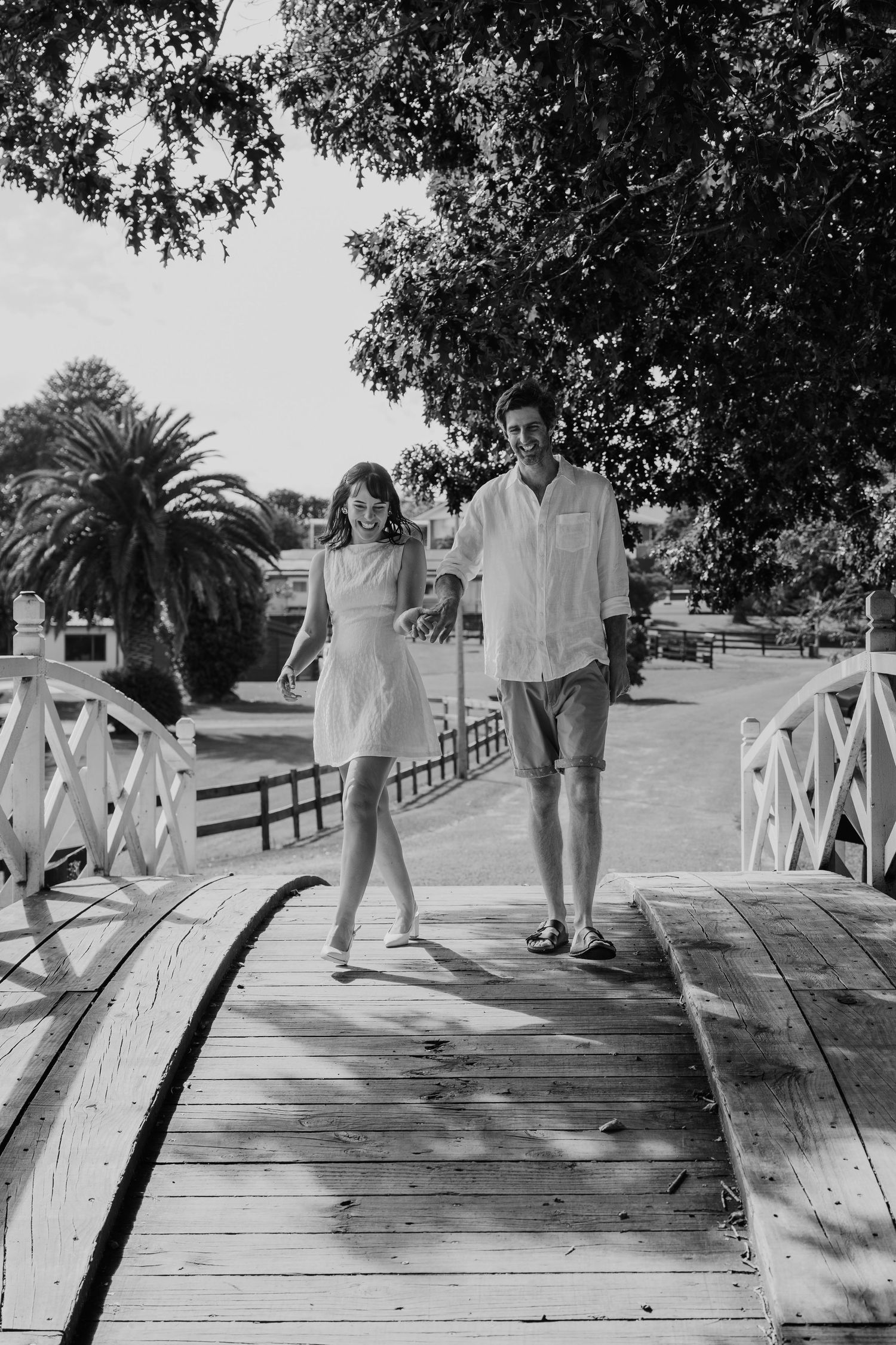 A couple walks hand in hand across a wooden bridge in a sunny park with palm trees and lush greenery in black and white. Elopement Tauranga