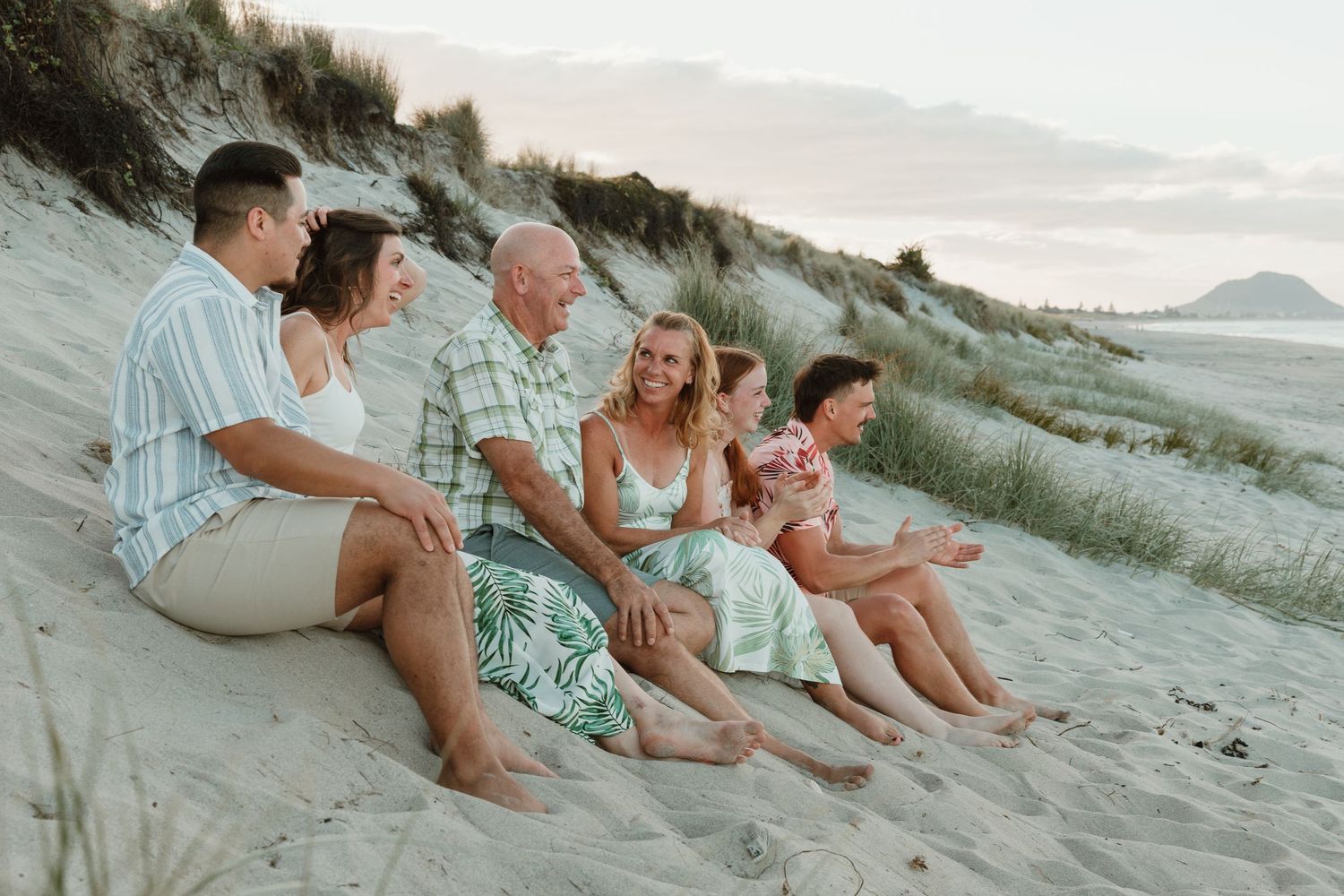 Family of five sitting together on sandy beach dunes at sunset, smiling and enjoying quality time outdoors.