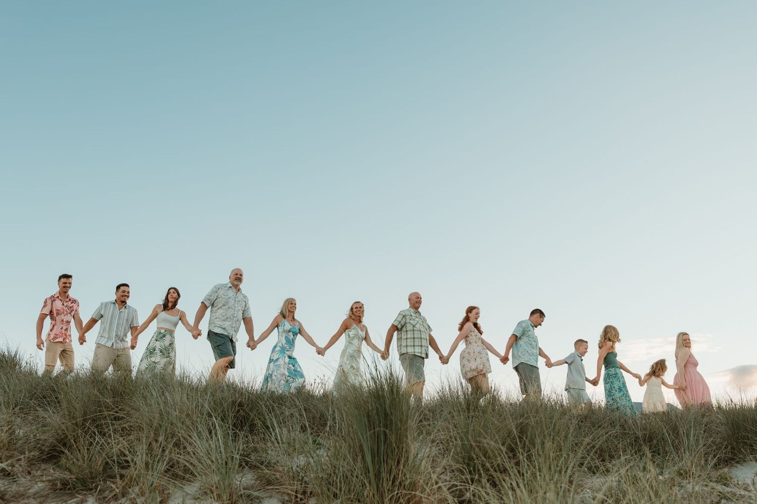 Large family holding hands in a line on sandy dunes at sunset, wearing coordinated pastel outfits. Extended Family photoshoot Tauranga