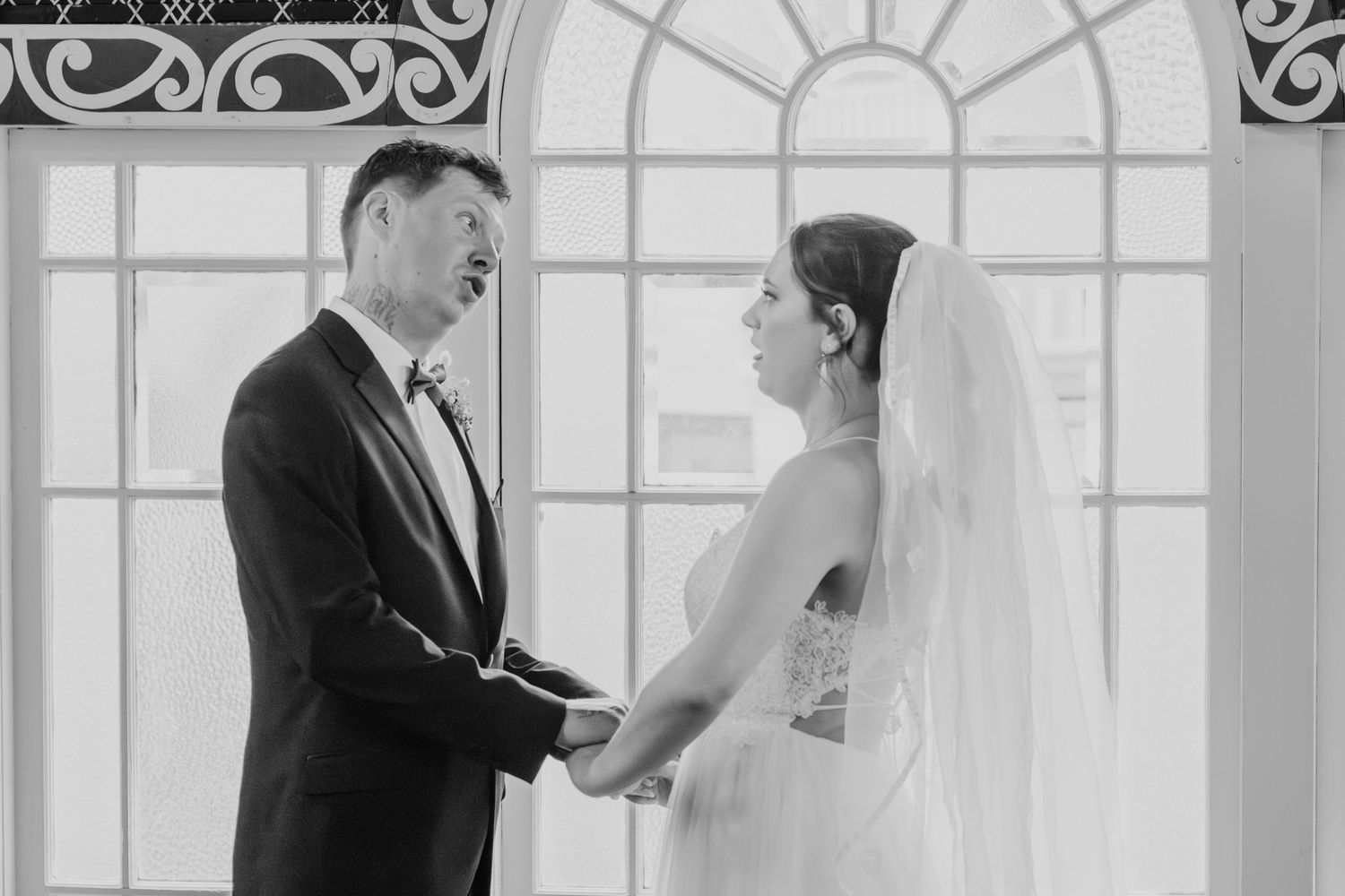 A black and white wedding photograph showing a couple holding hands in front of an ornate arched window.