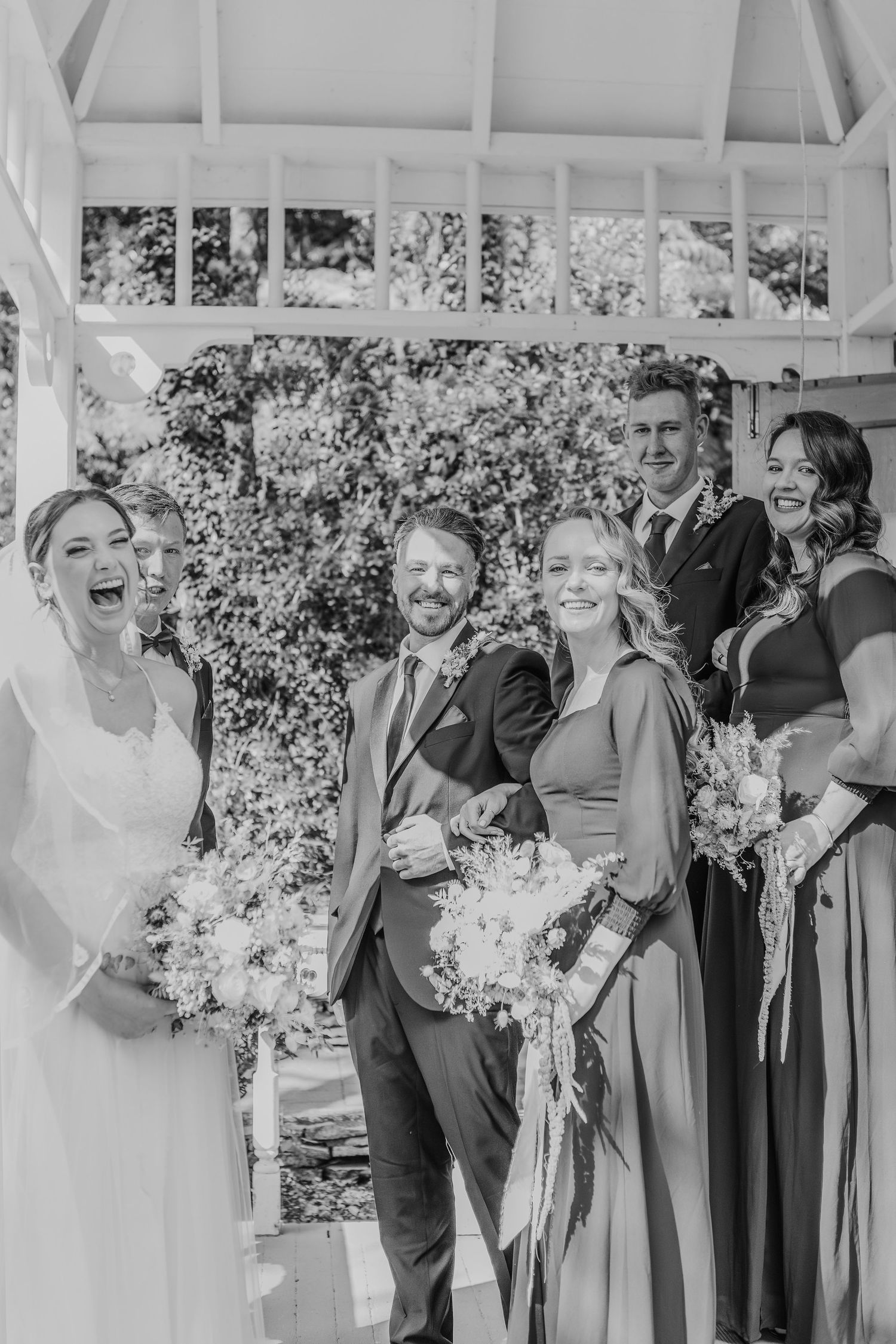 Black and white photo of bridal party laughing together during wedding ceremony in outdoor gazebo with floral bouquets.