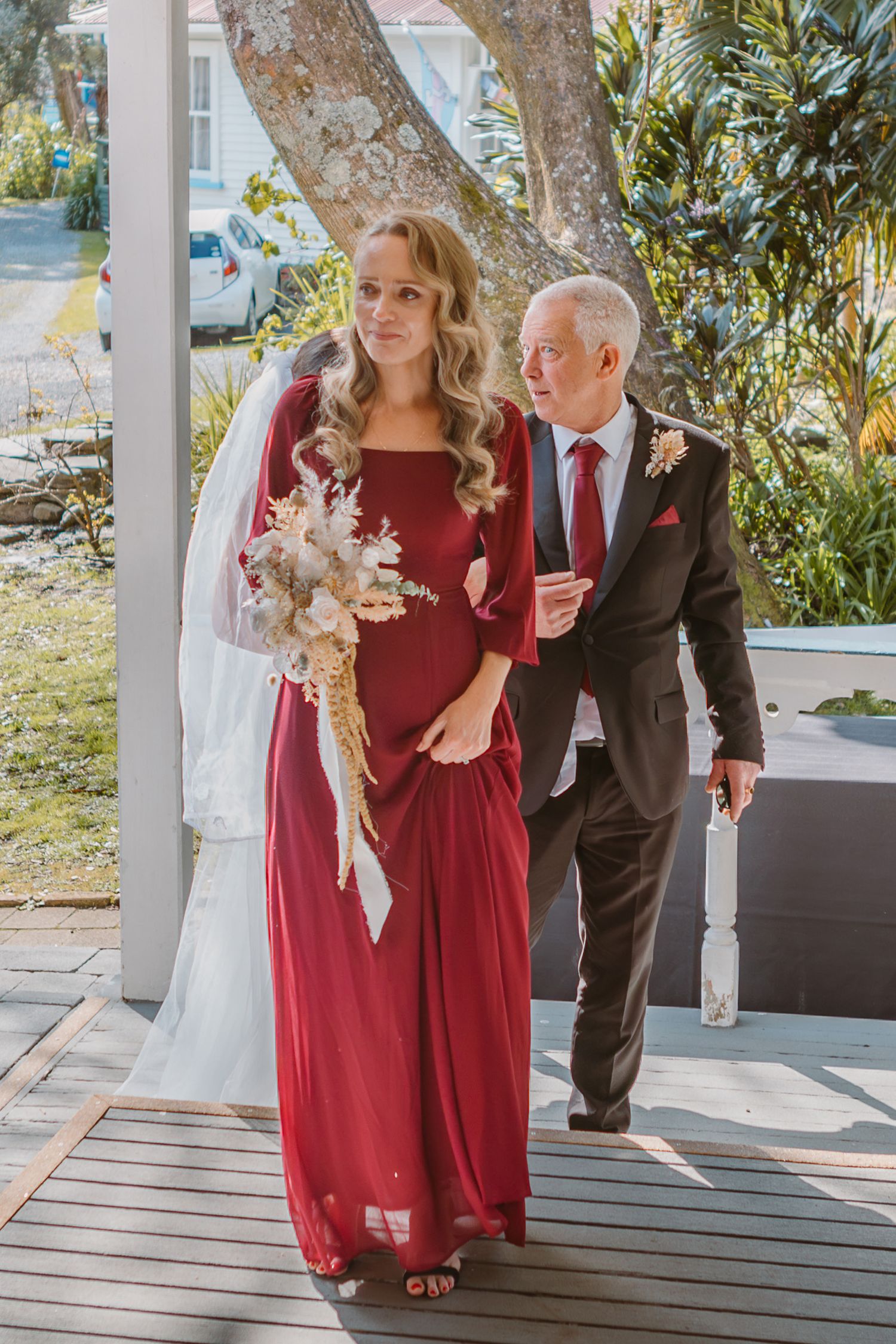 Two people walk together on a covered porch, one wearing a flowing burgundy dress and holding a floral bouquet.