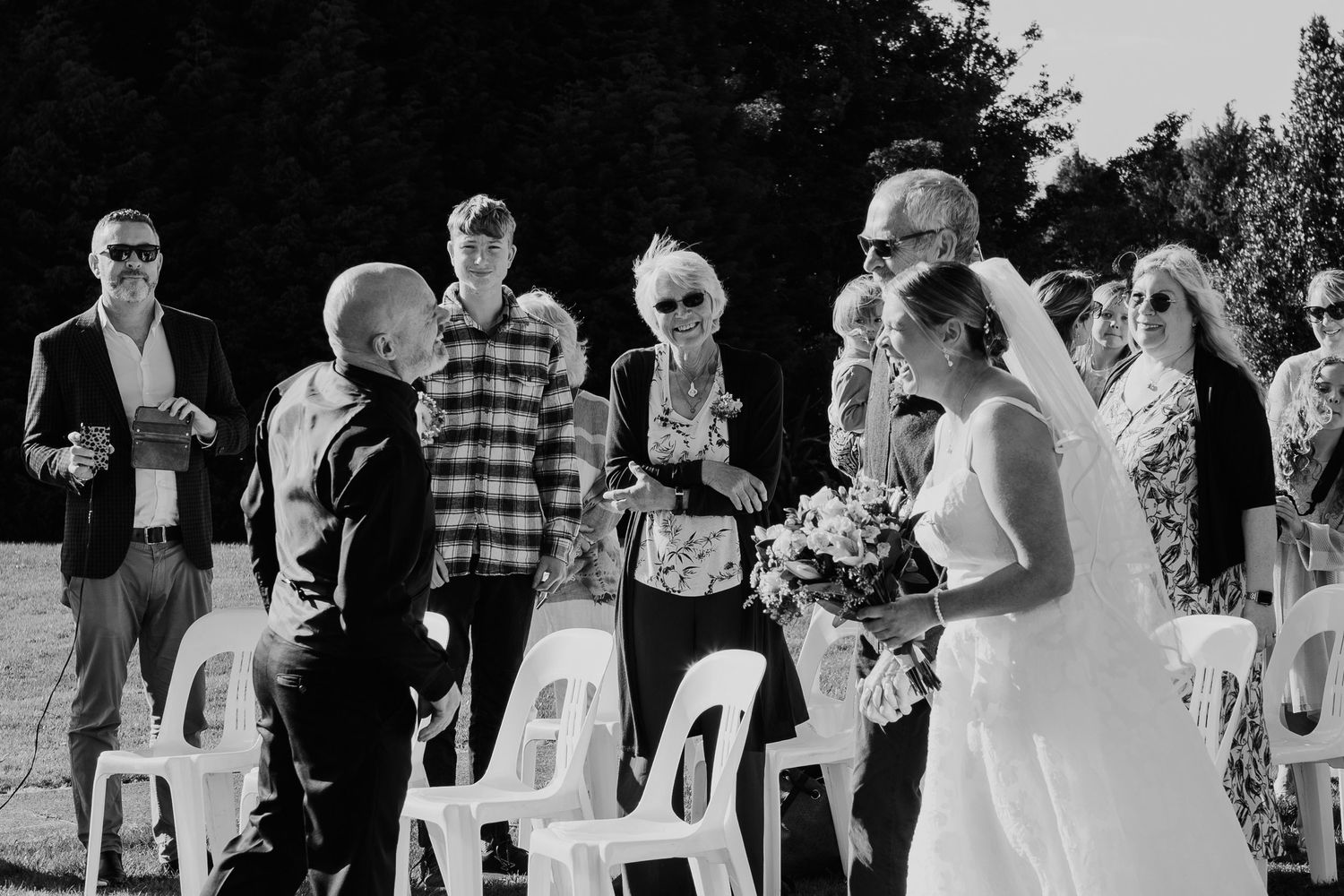 Black and white photograph of an outdoor wedding ceremony with guests seated in white chairs on a sunny day.