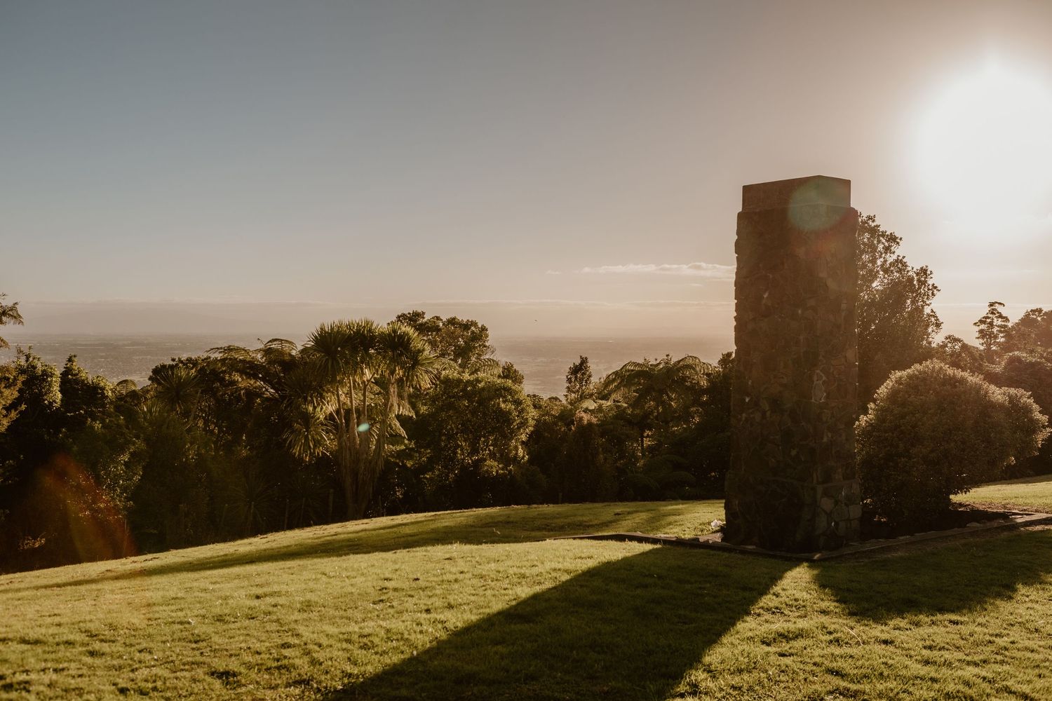 Sun casts golden light across a hilltop lawn with silhouetted trees and a tall stone structure against a hazy horizon.