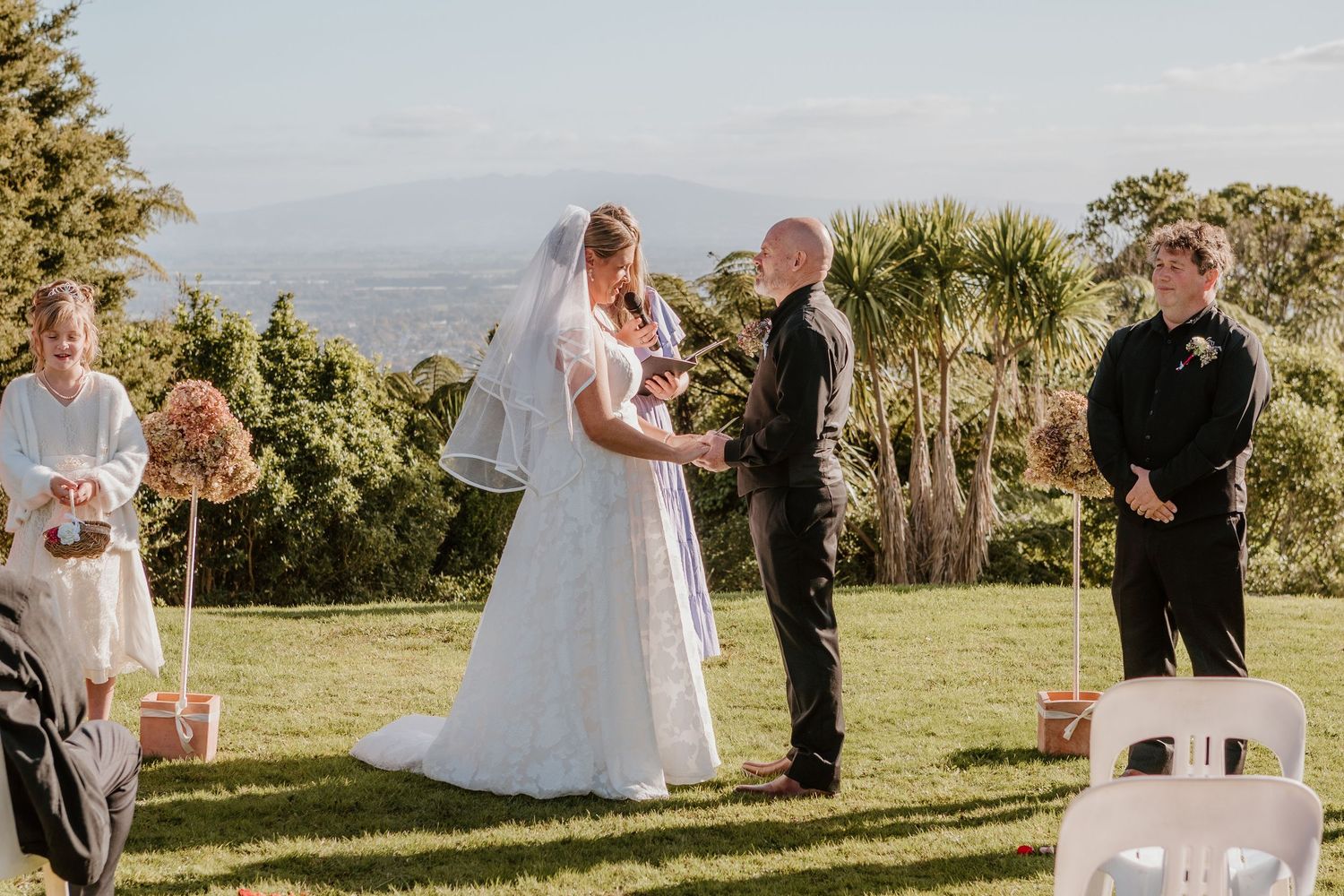 An outdoor wedding ceremony takes place on a hilltop venue with palm trees and mountain views in the background.