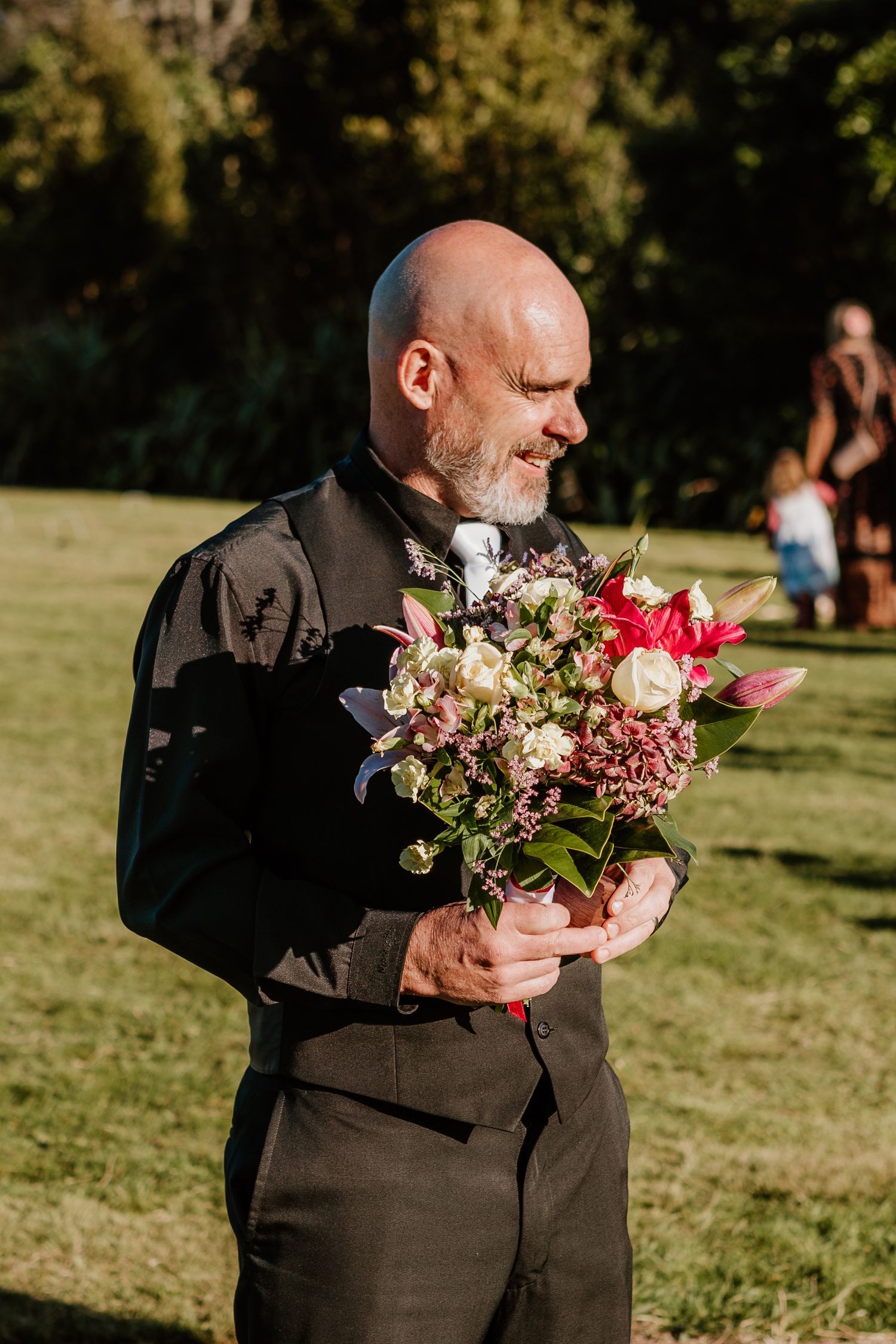 A person in a dark suit smells a beautiful bouquet of pink and red flowers in bright outdoor sunlight.