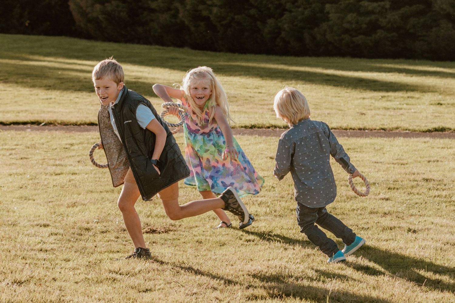 Three children run and play together in a grassy field on a sunny summer afternoon.
