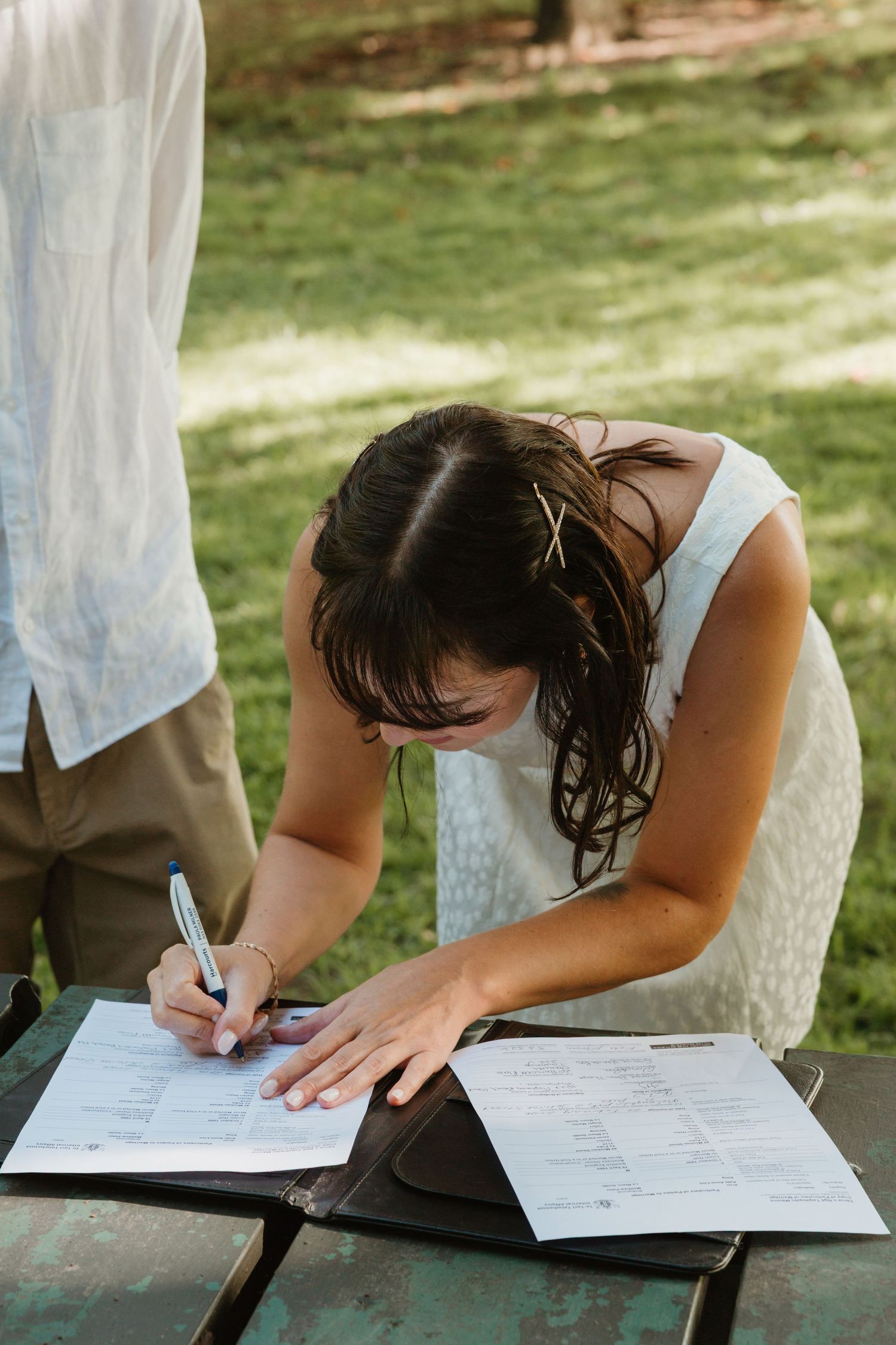 Bride in white dress signing marriage certificate at outdoor picnic table during wedding ceremony.