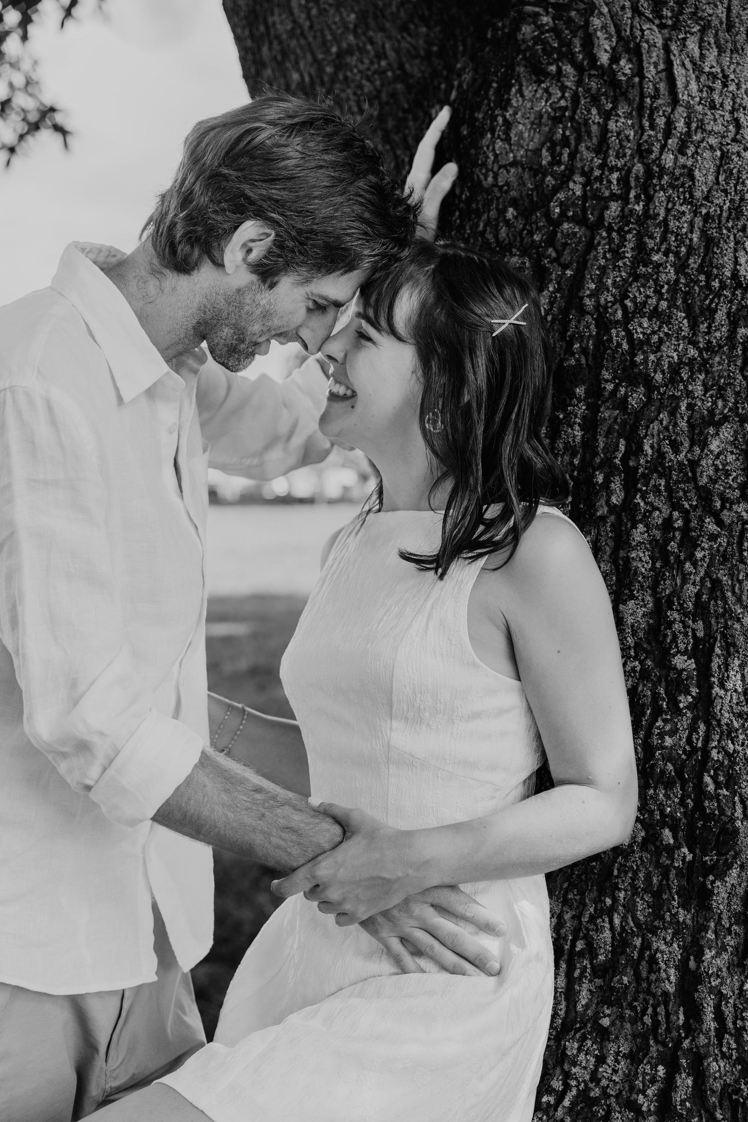 Black and white photo of a smiling couple leaning against a tree, sharing a tender romantic moment outdoors.