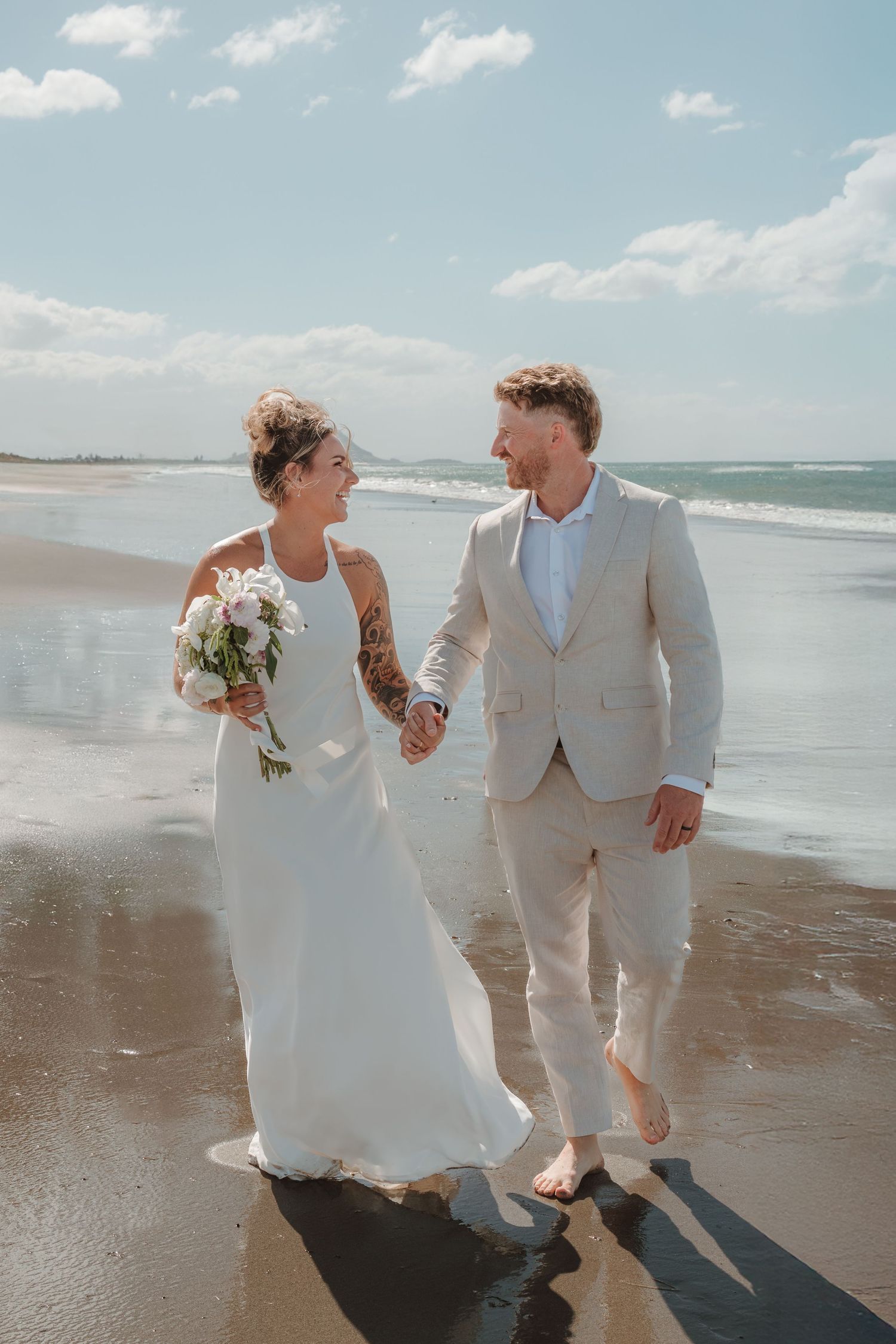 Bride and groom walking hand in hand on a sunny beach, bride holding a bouquet in a white gown, groom in a beige suit.