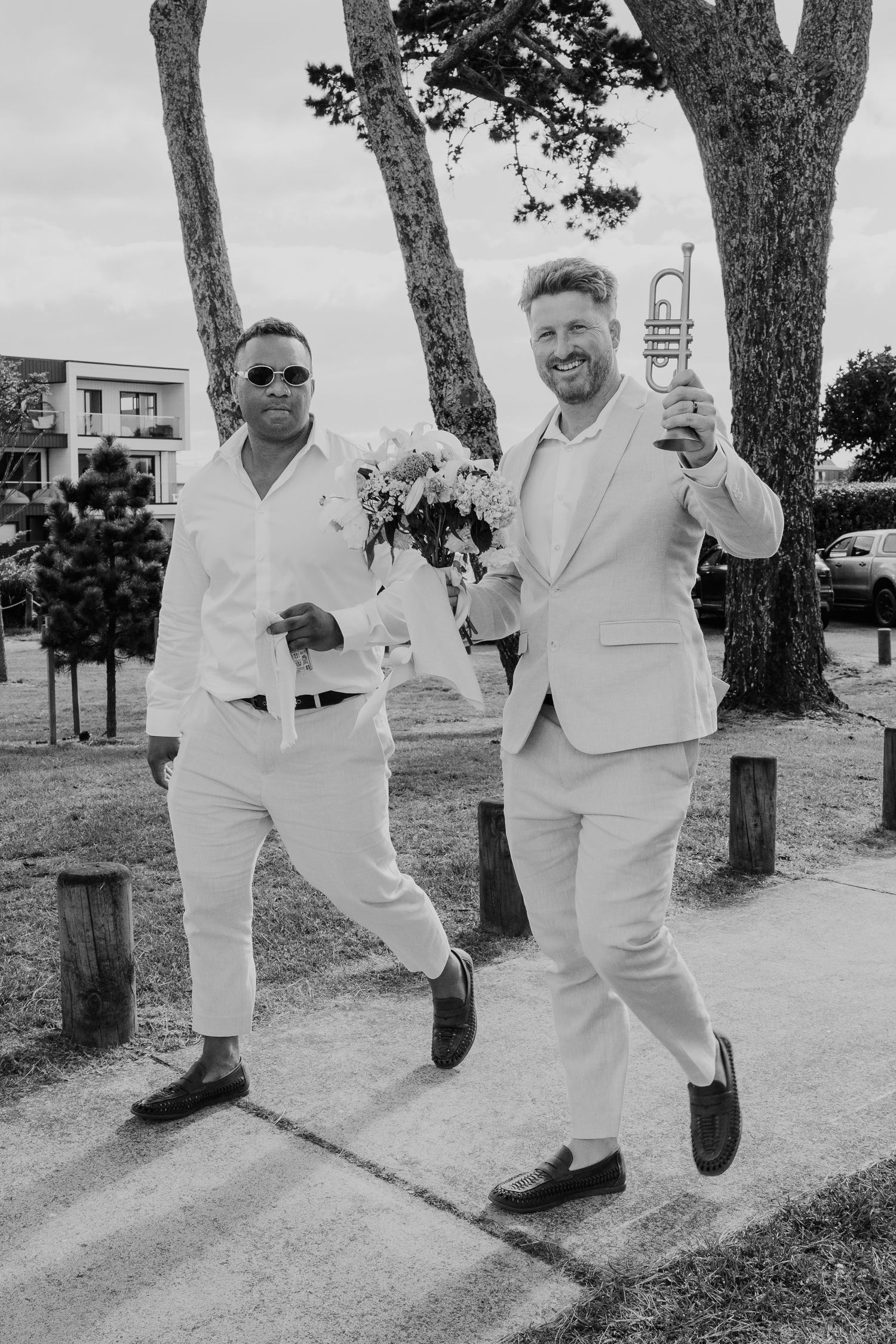 Two men in white suits celebrate outdoors, one holding flowers and one raising a trumpet, in a joyful black and white photo.