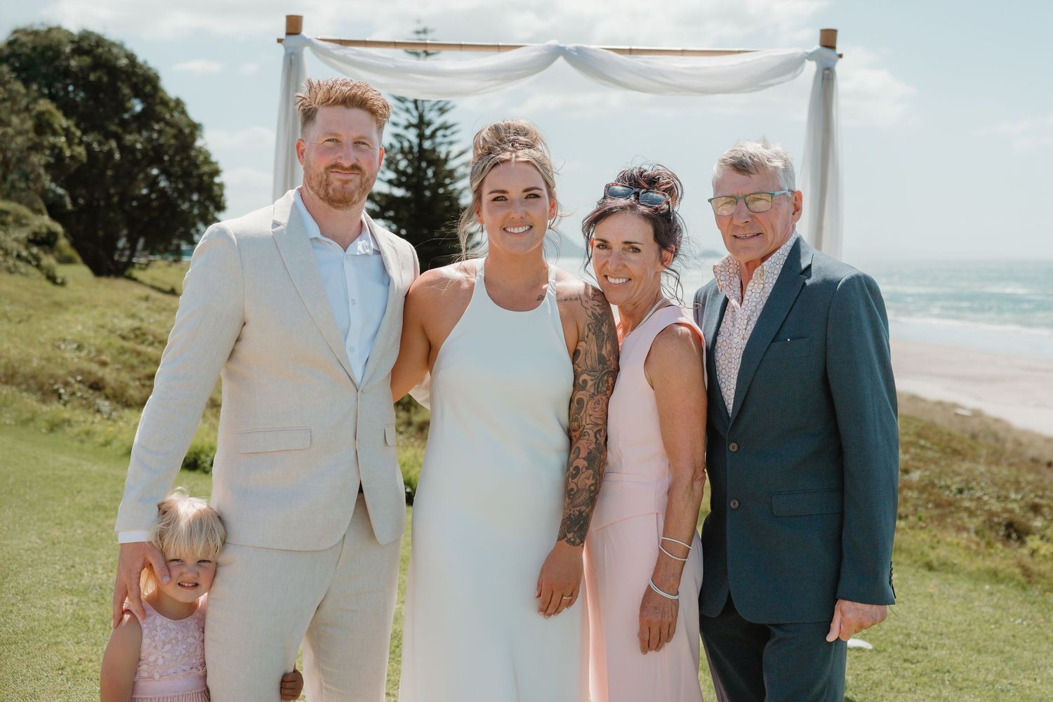 Wedding party of five posing outdoors near beach arch, bride in white dress with floral tattoos, guests in formal attire.