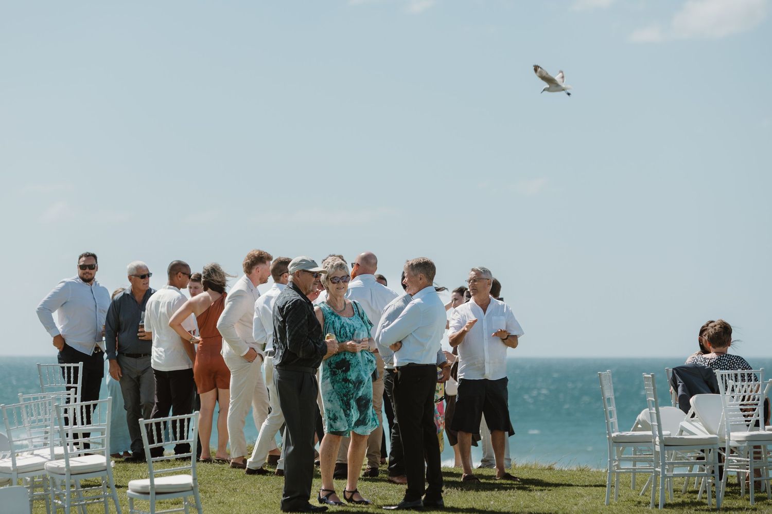 Guests mingling at an outdoor beachside wedding ceremony with white chairs and ocean views under a clear blue sky.