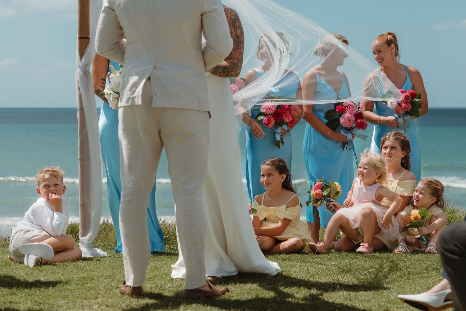 Beach wedding ceremony with bride and groom, bridesmaids in blue dresses, and children seated on grass by the ocean.