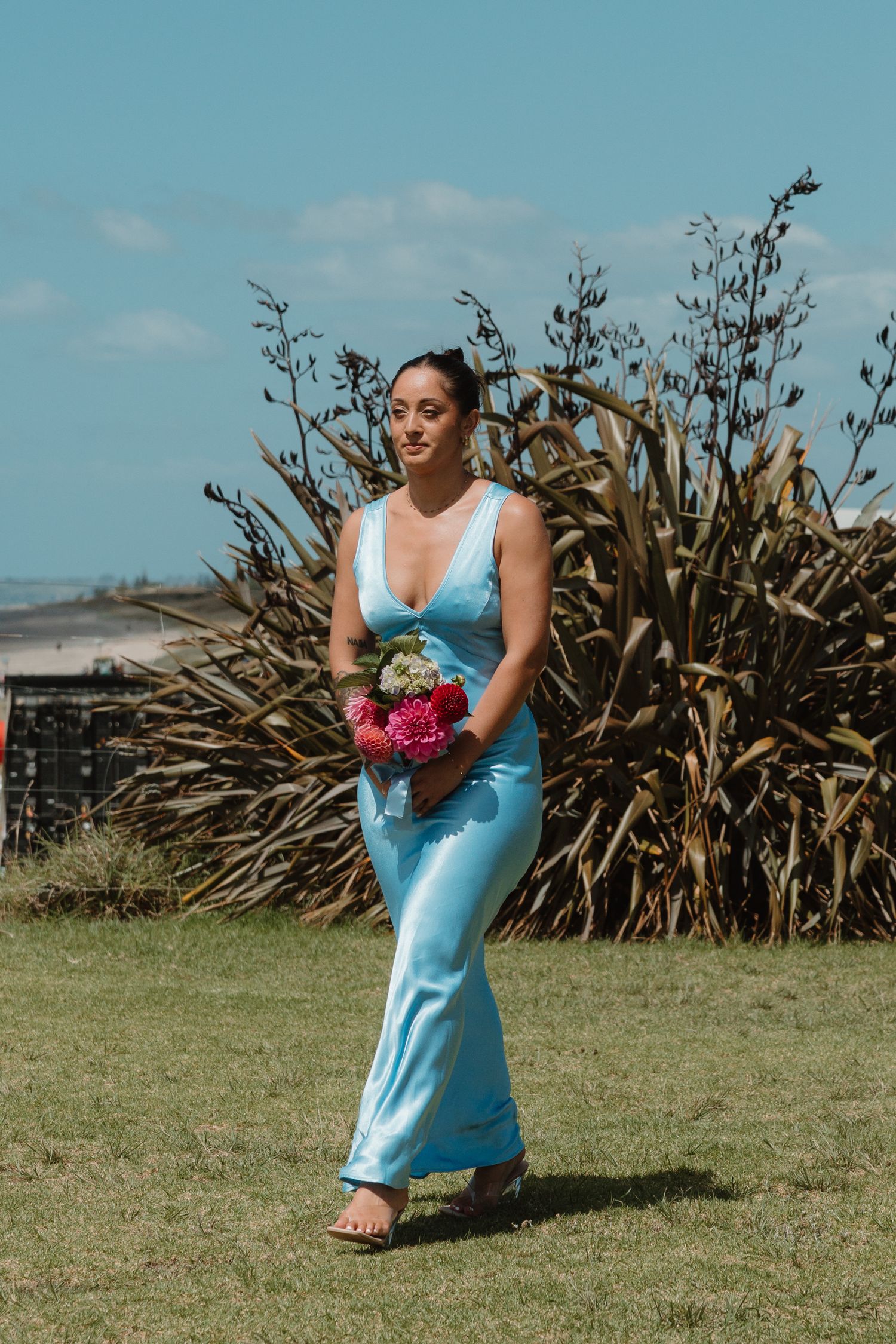 Woman in light blue satin gown holding pink bouquet, standing on grass with tropical plants and blue sky.
