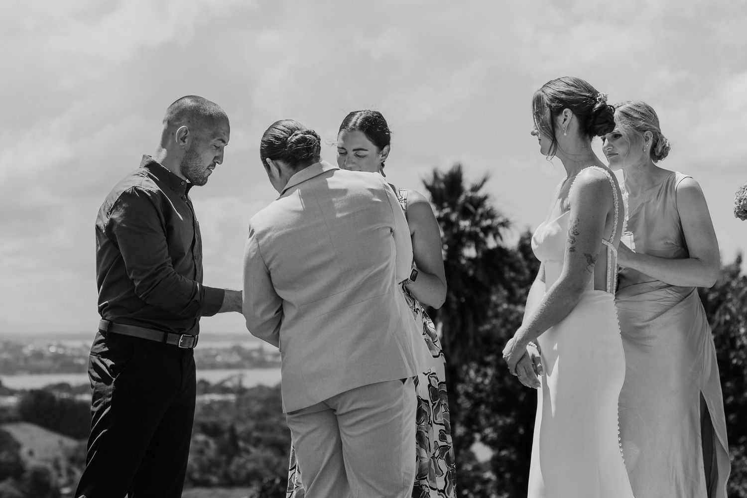 Black and white wedding ceremony photo of couple exchanging vows outdoors with officiant and bridesmaid on a scenic hilltop.