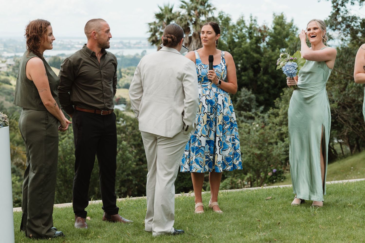 Wedding party gathered on hilltop during outdoor ceremony, bridesmaid in mint dress holds blue bouquet.