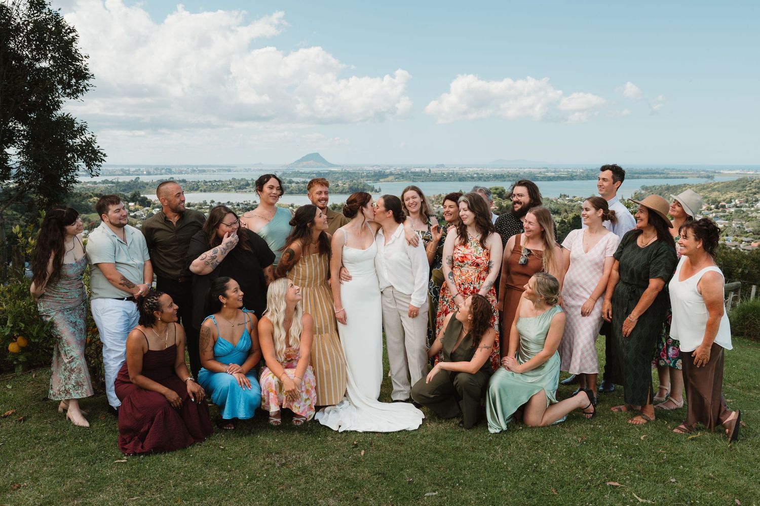 Wedding group photo outdoors with scenic coastal views, featuring bride and groom surrounded by guests on a green hillside.