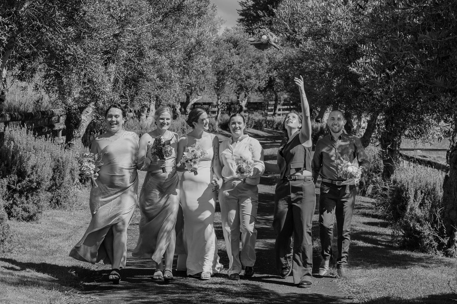 Black and white photo of a joyful wedding party walking outdoors among trees, with bride and bridesmaids celebrating.