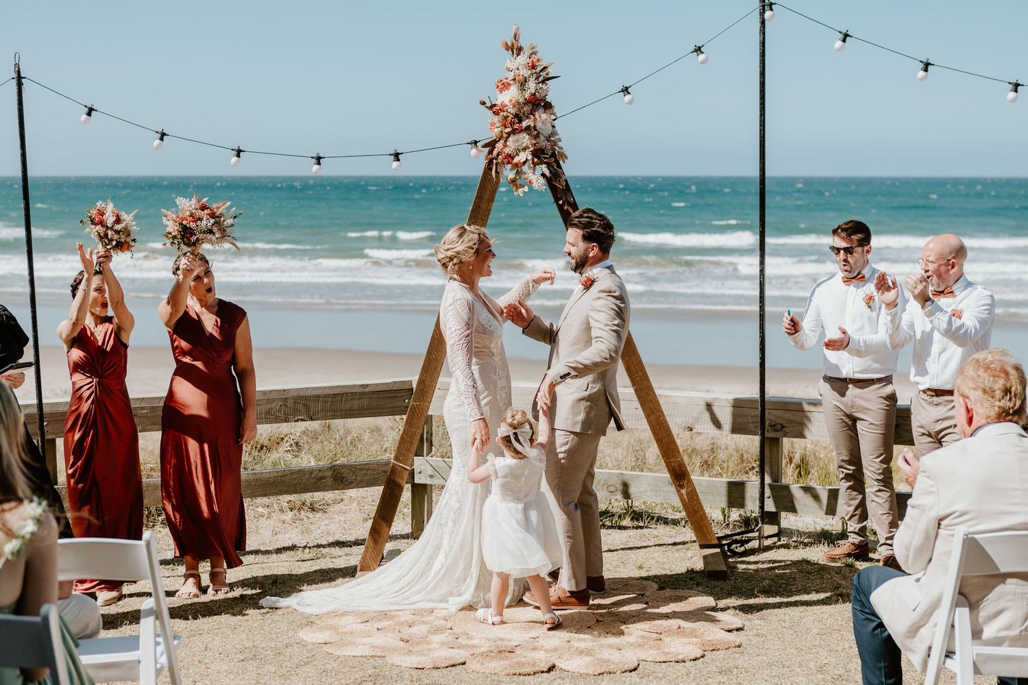 Beachside wedding ceremony under a wooden arch with string lights and ocean backdrop. Papamoa Surf Lifesaving Club Wedding