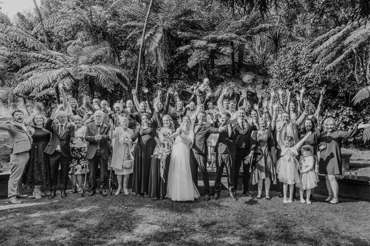 Wedding guests and family members pose for a celebratory group photo in a tropical garden setting.