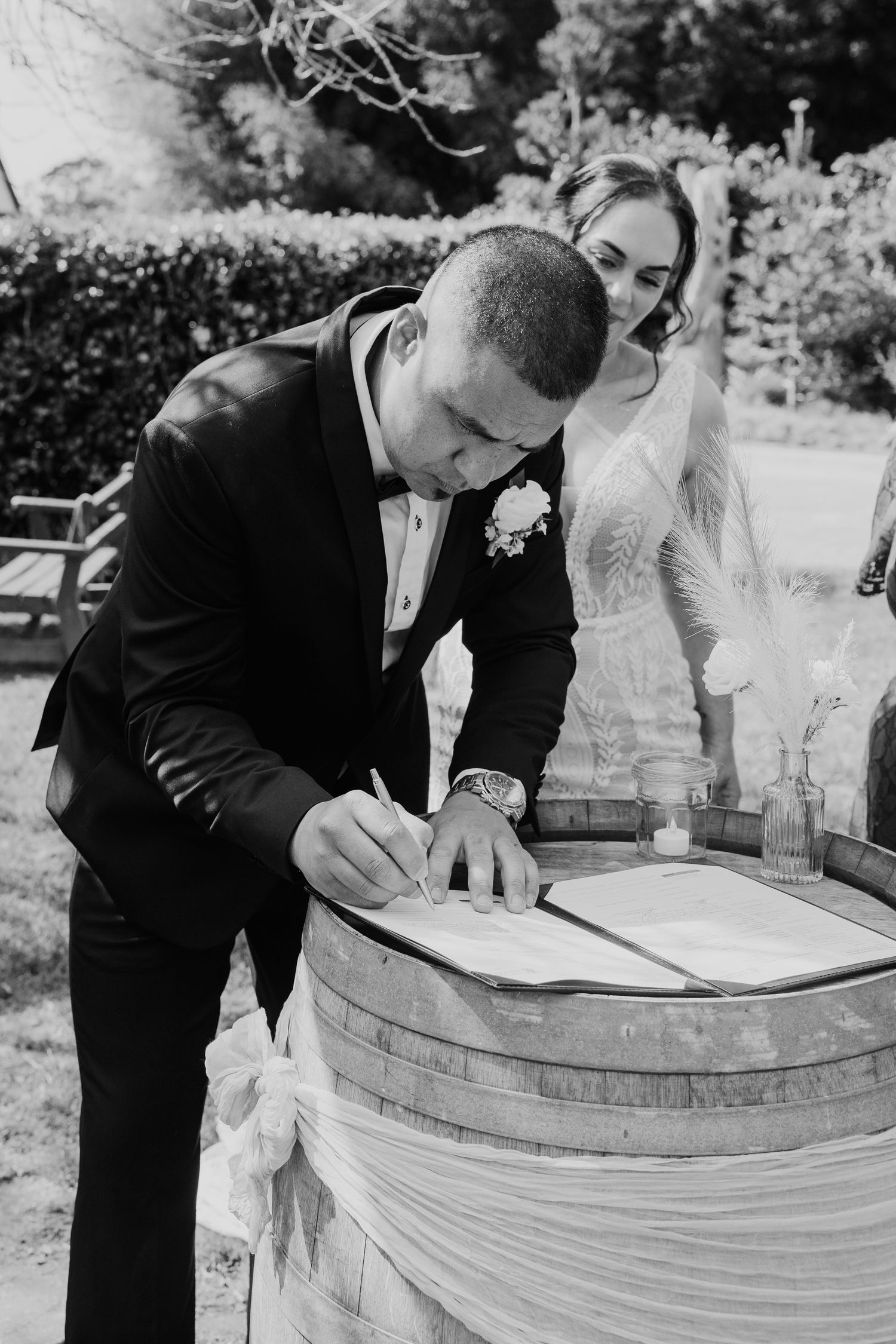 A series of black and white photos showing a couple signing their marriage license on a wine barrel at an outdoor wedding.