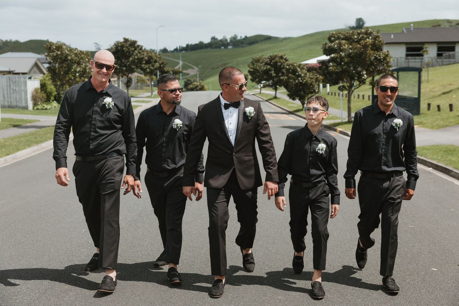 Five people in formal black attire walking together down a paved road with green hills in the background.