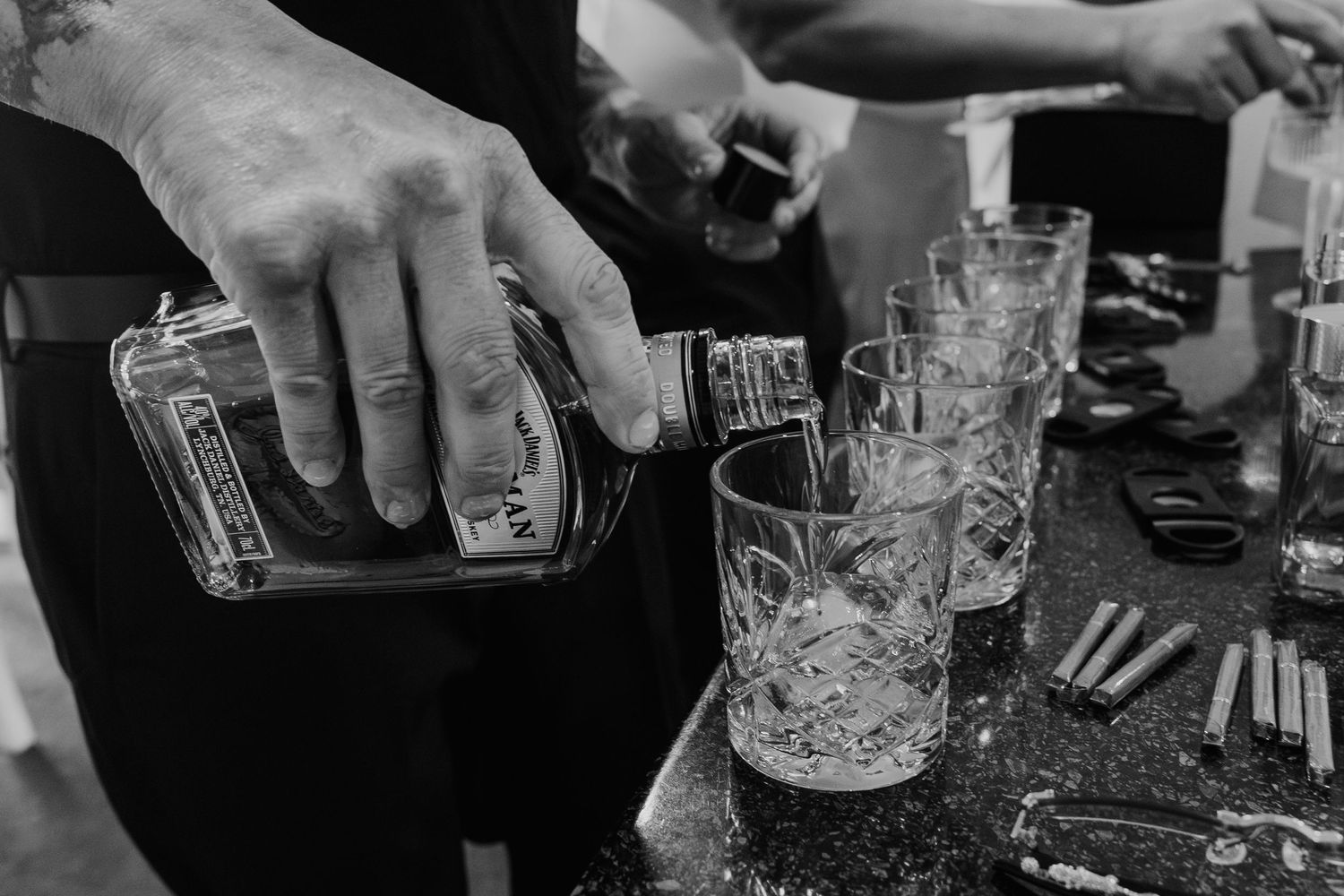 Black and white photo showing hands pouring drinks at a bar with multiple glasses visible in a row.