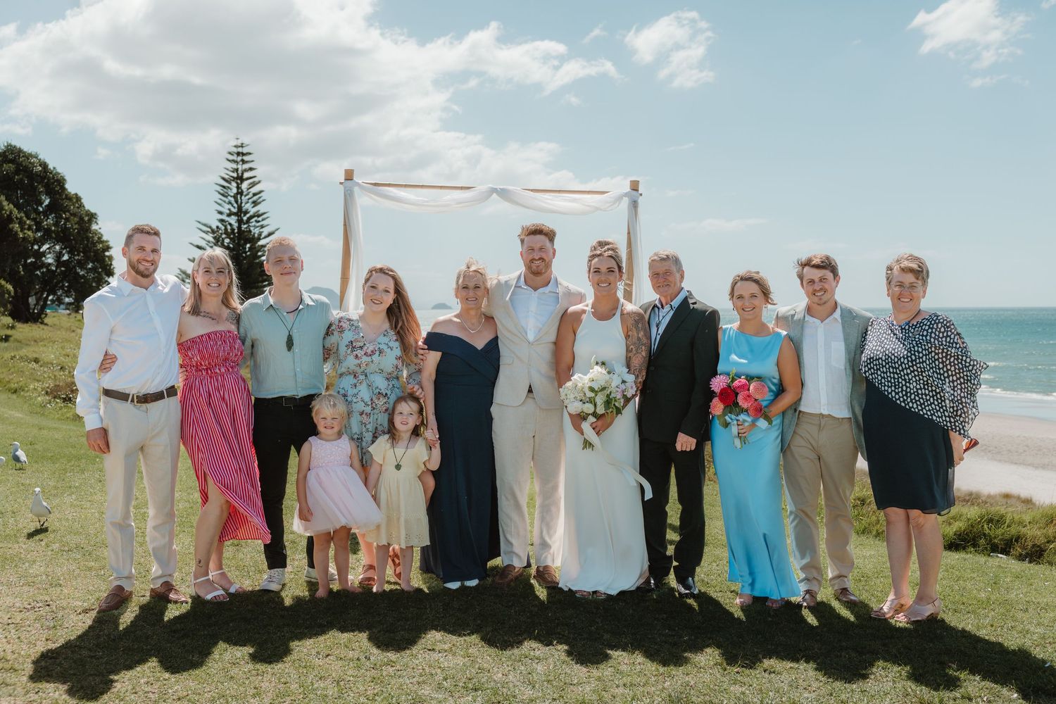 Wedding party posing outdoors near ocean with arch backdrop on sunny day.