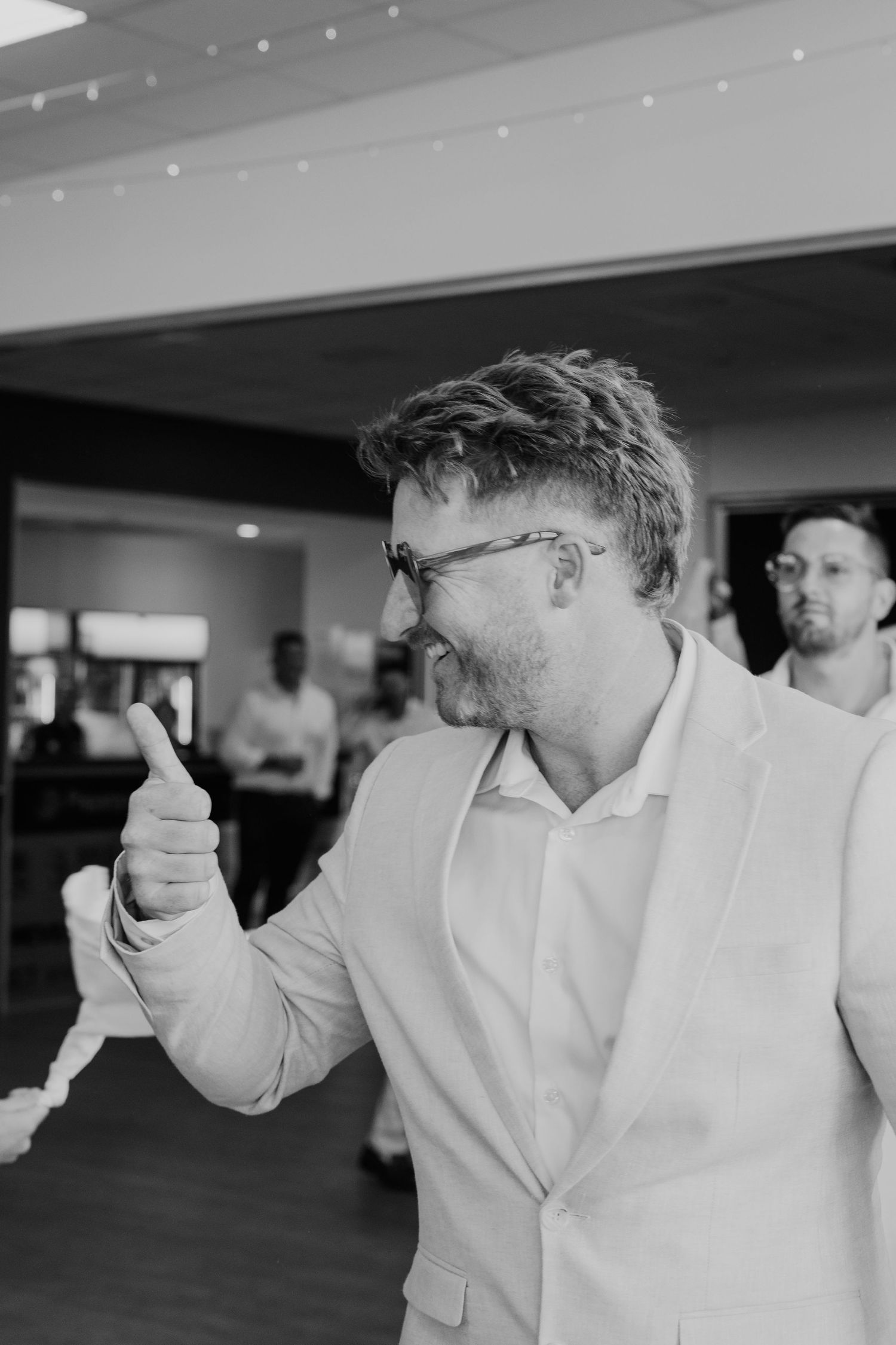 Black and white photo of a smiling man in a suit giving a thumbs up at a wedding reception.