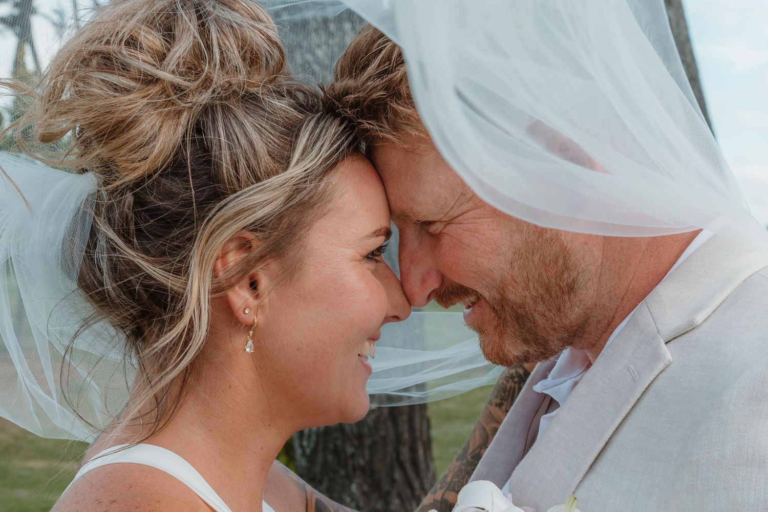 Bride and groom touching foreheads under a flowing veil, sharing an intimate romantic moment on their wedding day.