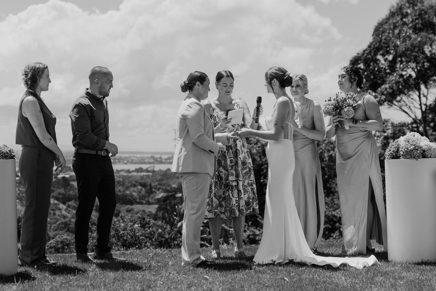 Black and white outdoor wedding ceremony with couple exchanging vows surrounded by guests on a scenic hilltop.