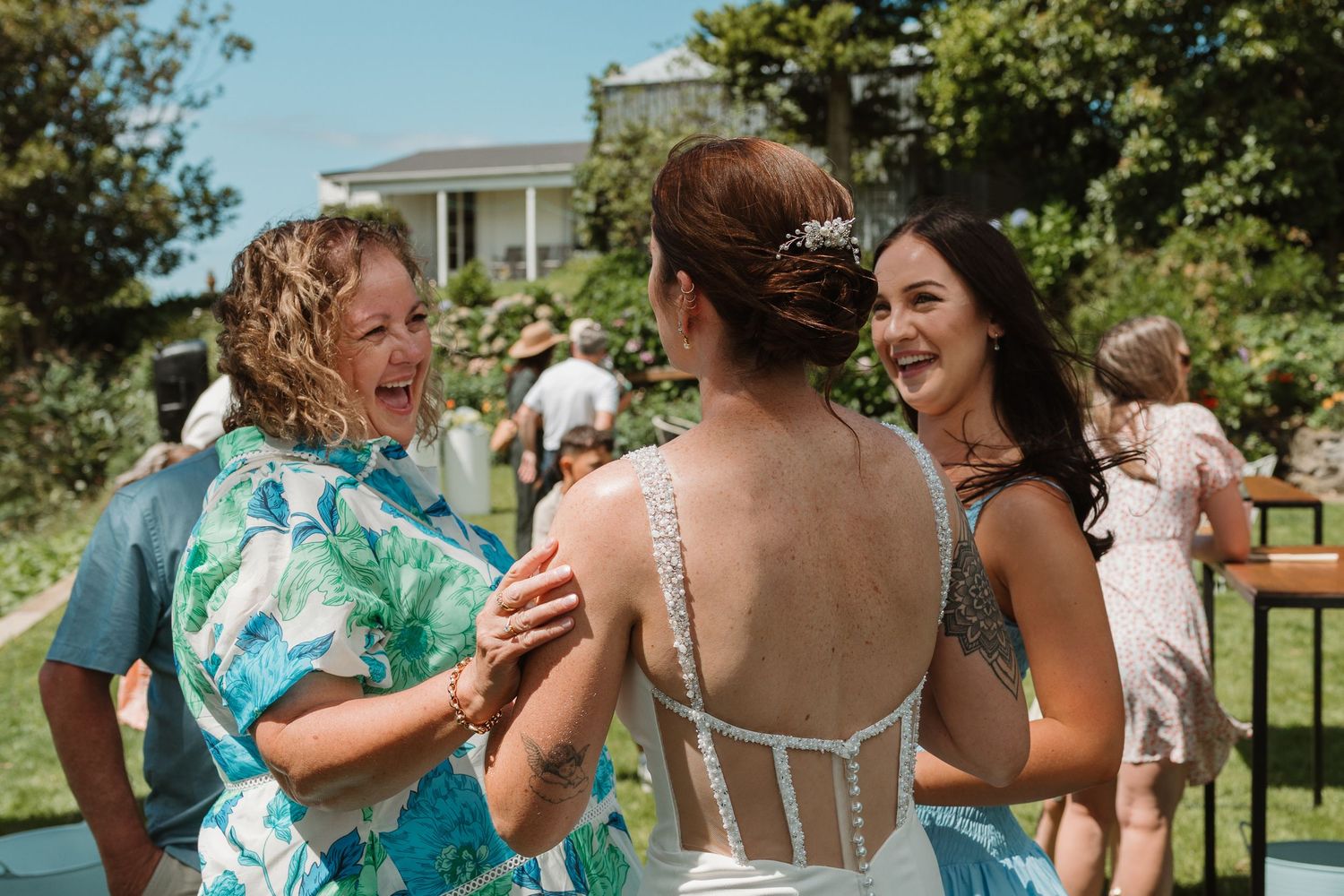 Bride in white gown with tiara laughing with two female guests at outdoor wedding reception.