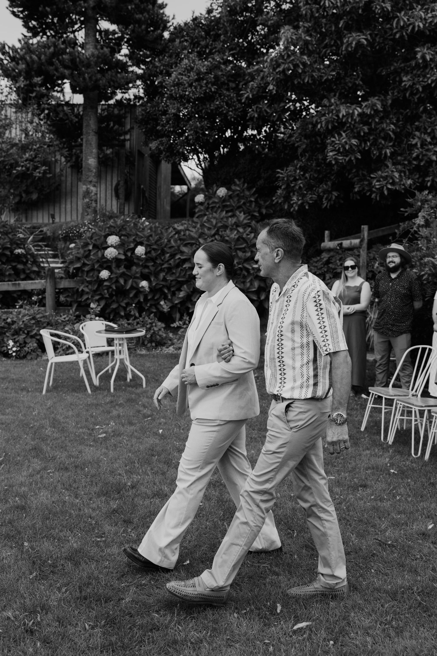 Black and white photo of two people walking arm in arm across a lawn at an outdoor garden party.