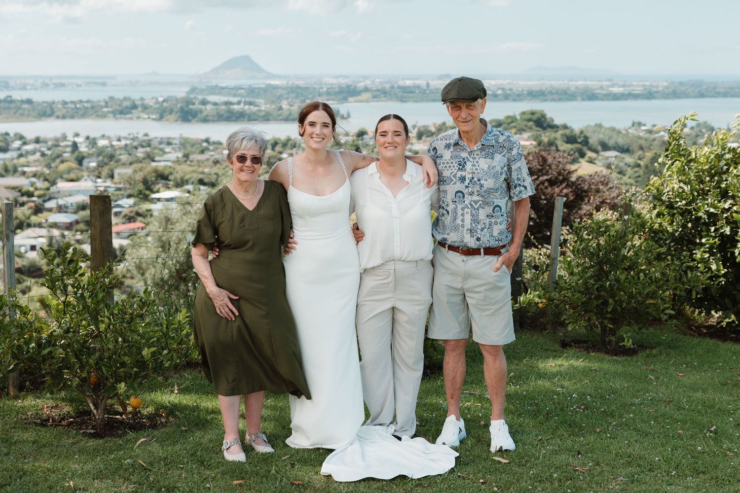 Bride in white gown poses with three guests on green hillside with scenic coastal town and mountain view.