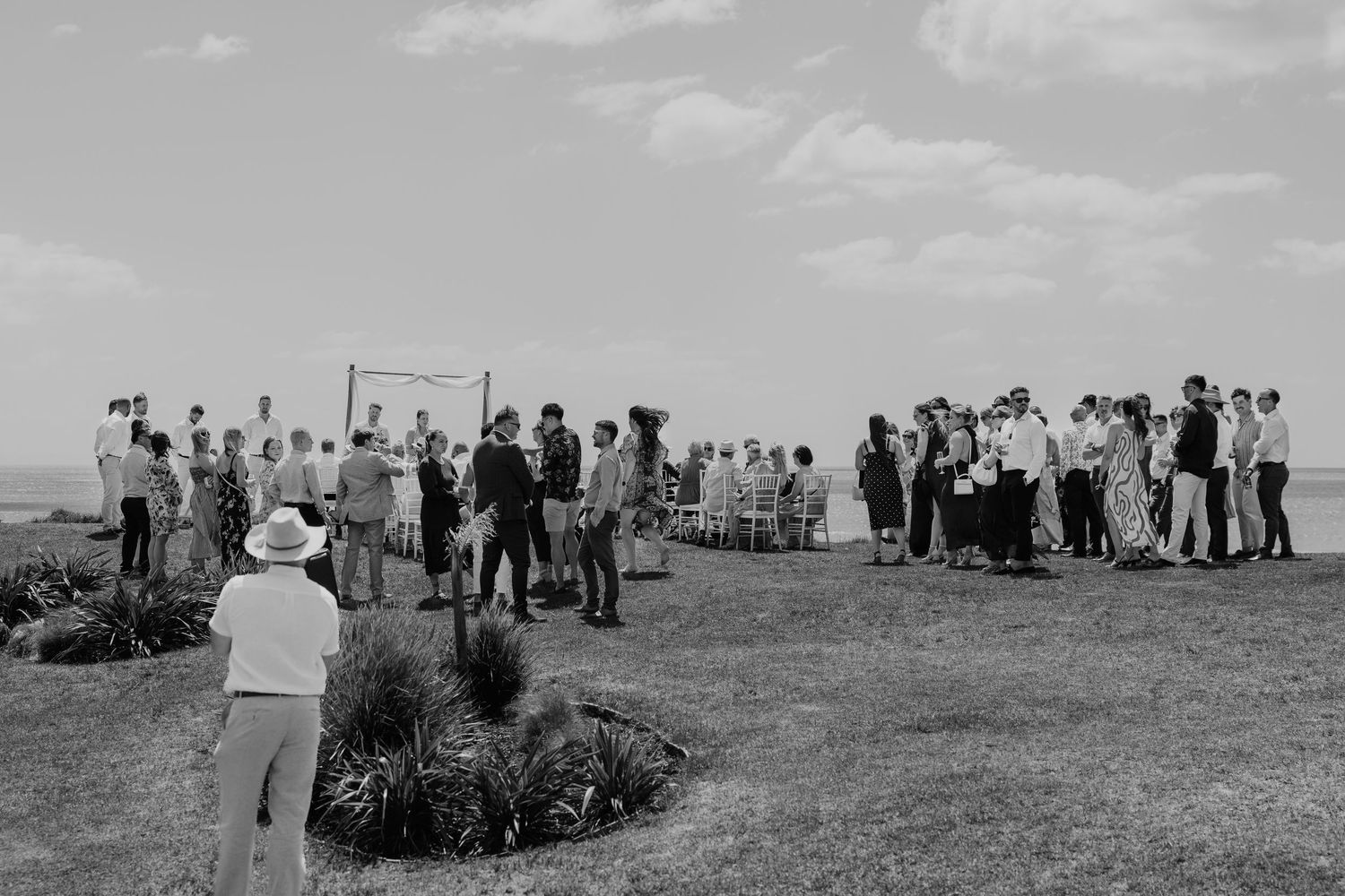 Black and white outdoor wedding ceremony with guests gathered on a grassy coastal lawn under a rustic arch.