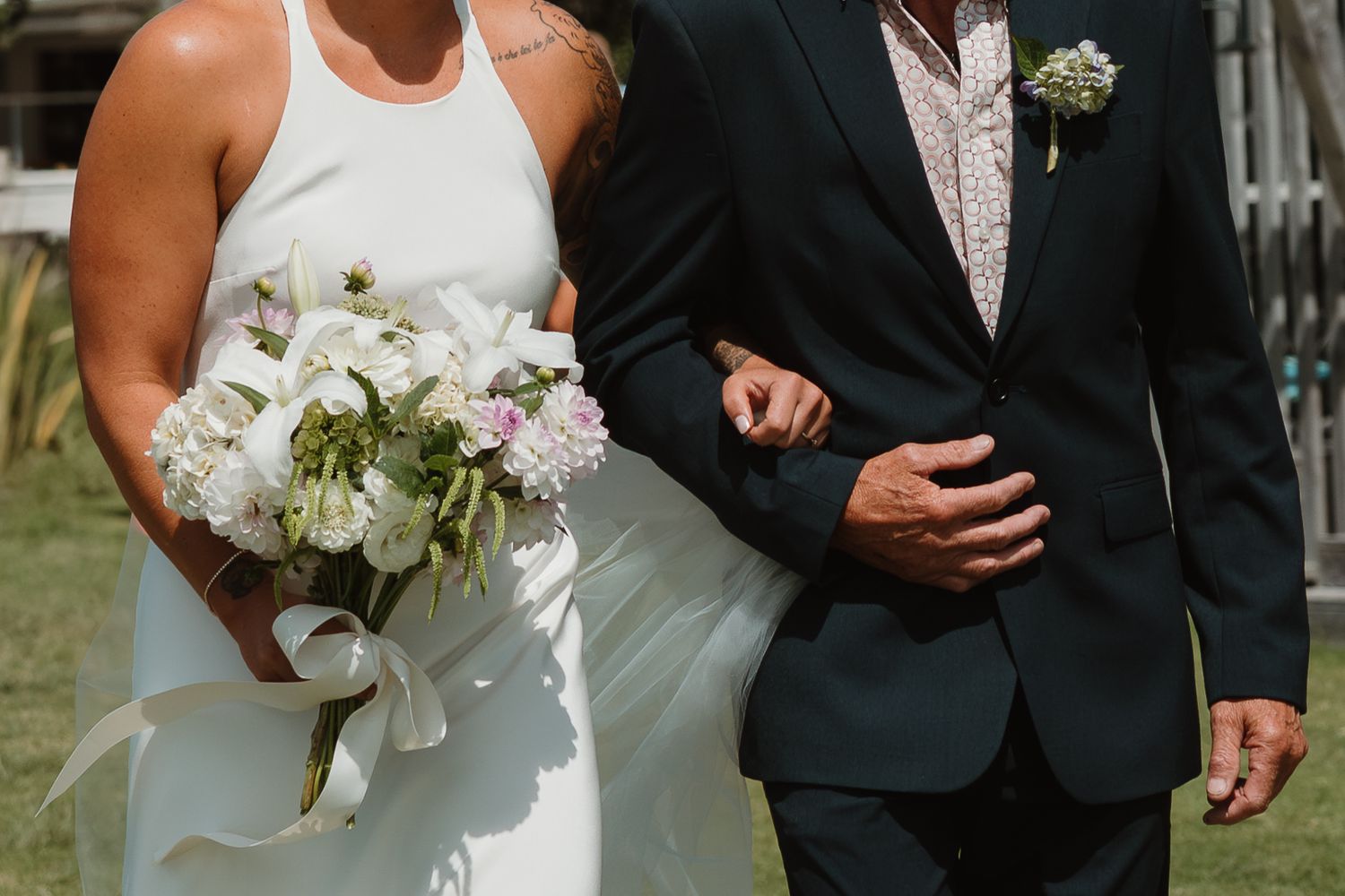 Bride in white dress holding bouquet walks arm-in-arm with groom in dark suit at outdoor wedding ceremony.
