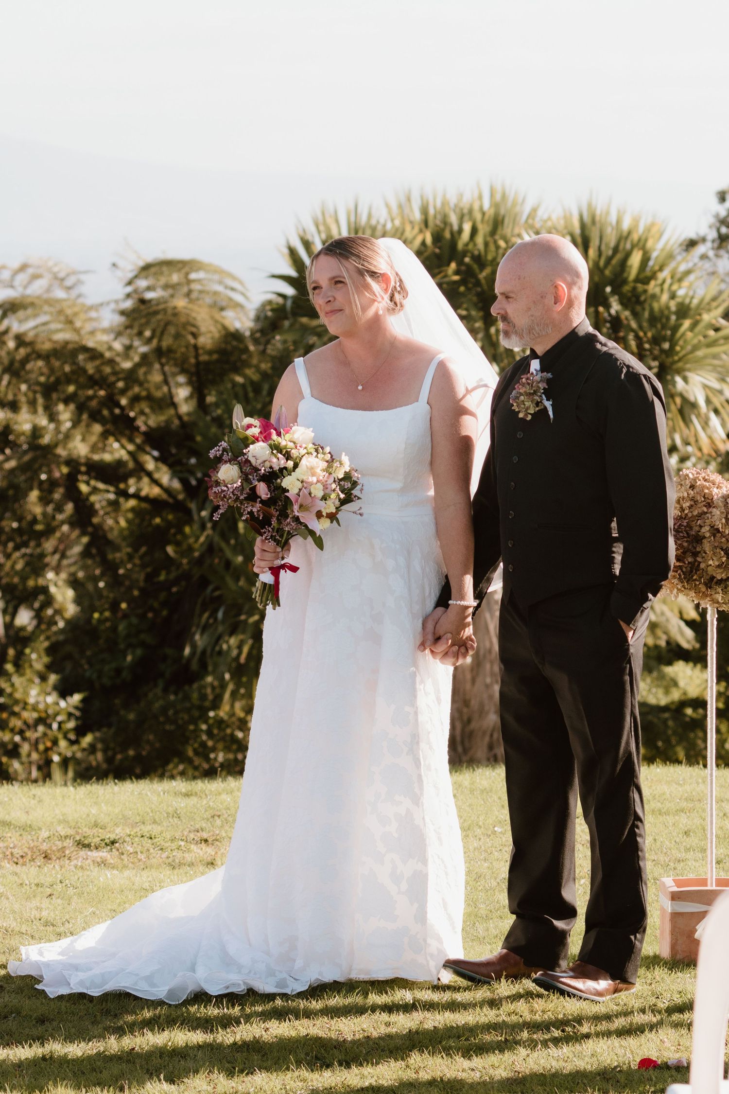 A bride in a white wedding gown holding a pink bouquet stands with a person in black attire outdoors near tropical foliage.