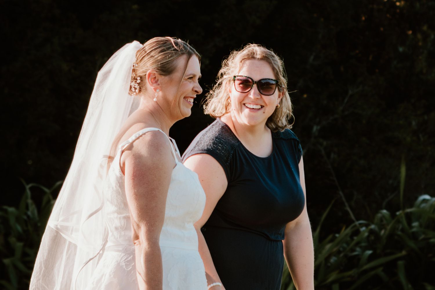 A bride in a white wedding gown shares a candid moment with a woman in a navy blue dress outdoors.